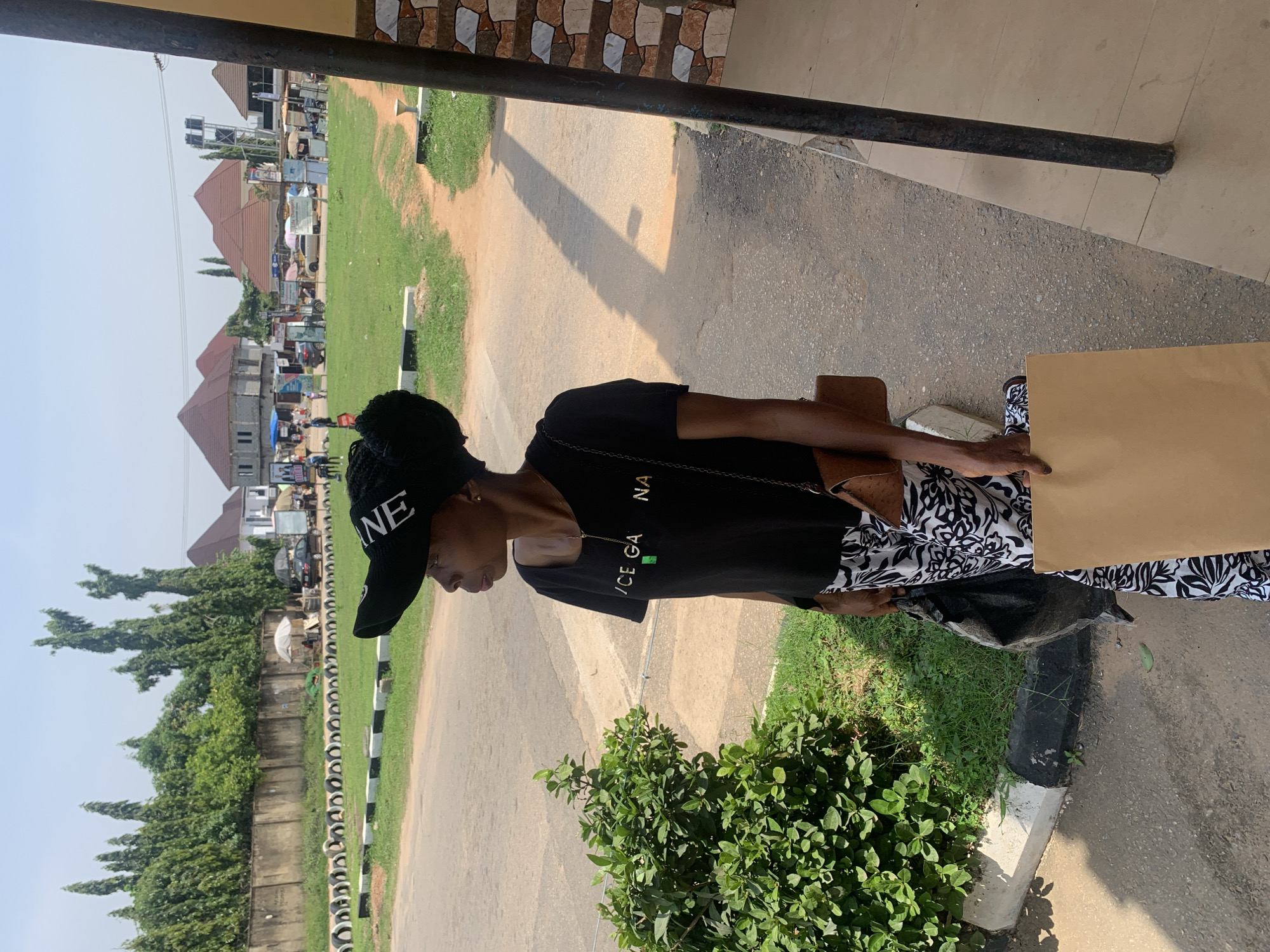 A young Black woman stands outdoors, looking to her left. She wears a black t-shirt with the words "ICE GANA" in white letters, a black baseball cap, and patterned black and white pants. She carries a brown purse on her right shoulder and a light brown paper bag in her right hand. A black plastic bag is held in her left hand. She is standing on a sidewalk, with a green bush to her left and a paved road and grass in the background. Buildings and trees are visible in the distance. The scene appears to be in a commercial area, possibly a marketplace, with several stalls and vehicles in the background, suggestive of a location within Abuja, Nigeria, given the architecture and vegetation. The lighting indicates it is daytime, with clear skies.