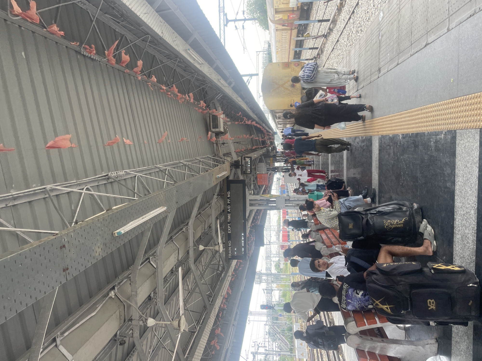 The image depicts a busy train station platform in Pathankot, India. A covered walkway extends along the platform, supported by metal structures and illuminated by fluorescent lights. Red pennants are strung along the underside of the roof, possibly for decoration or to ward off birds. Various people are visible on the platform, some sitting on benches and others standing and walking. Many are carrying luggage. Two yellow trains are parked on adjacent tracks, with one train appearing to be at the platform. The platform itself is made of concrete with a yellow tactile paving strip along the edge. A digital display board above the platform indicates "TRAIN NO. EXPT. TIME A/D PF. NO.". In the background, more tracks and railway infrastructure are visible, along with some greenery and buildings. The overall atmosphere appears to be one of transit and everyday life at a public transportation hub.