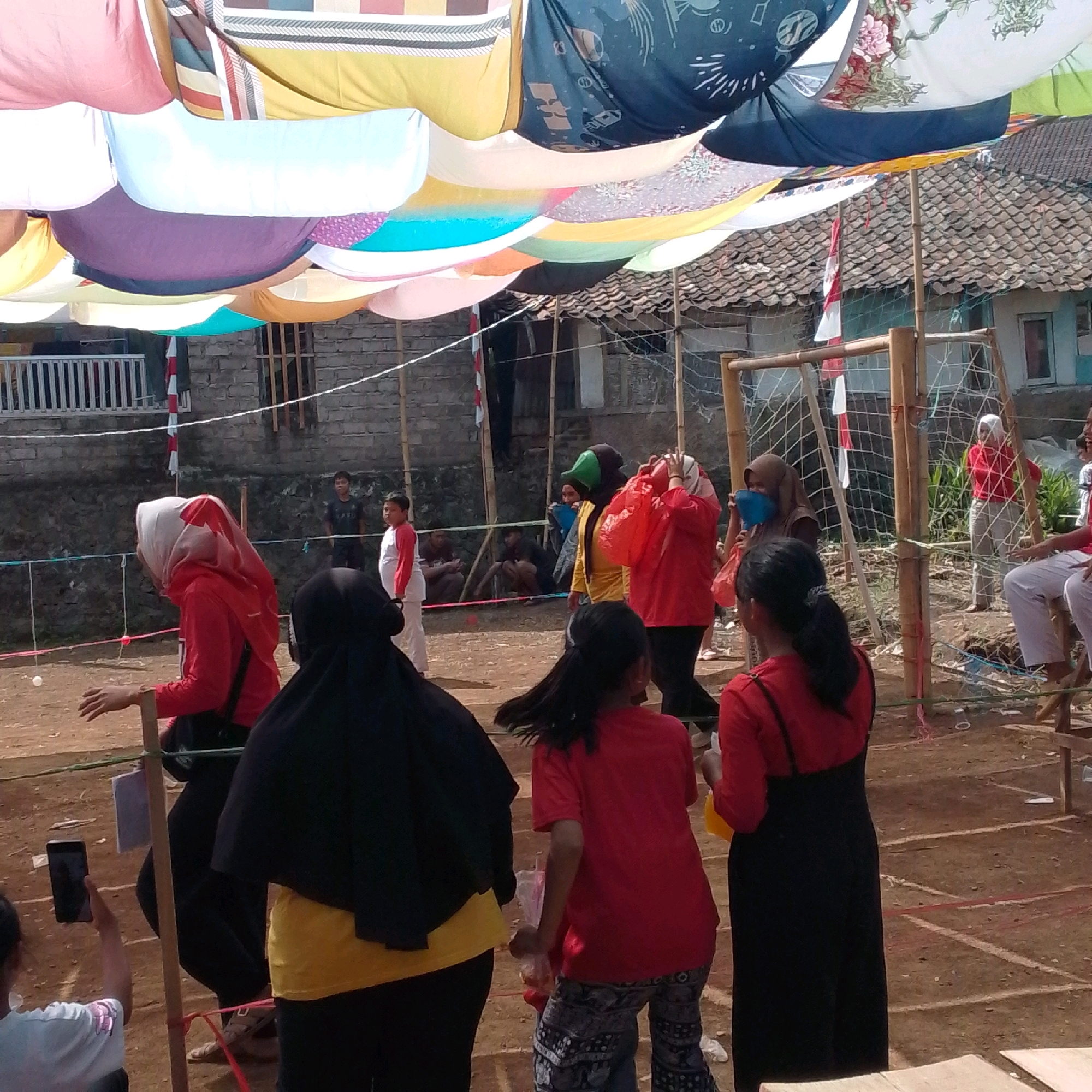 In Cilaku, Indonesia, a daytime outdoor event is captured, possibly a community gathering or sports competition. A makeshift canopy of colorful fabrics is suspended overhead, providing shade. In the foreground, a person in a red hijab and red long-sleeved shirt is seated, possibly in a wheelchair, with their left hand extended. Another person in a black hijab and yellow undershirt is standing next to them. Behind them, several other individuals are visible, including some in red shirts and one in a yellow jacket. A bamboo structure with netting, resembling a goal or net, is in the midground. People are gathered around this area, and a few children can be seen in the background. The ground is dirt, marked with white lines, suggesting an athletic field or activity area. The architectural style of the buildings in the background indicates a rural or suburban setting.