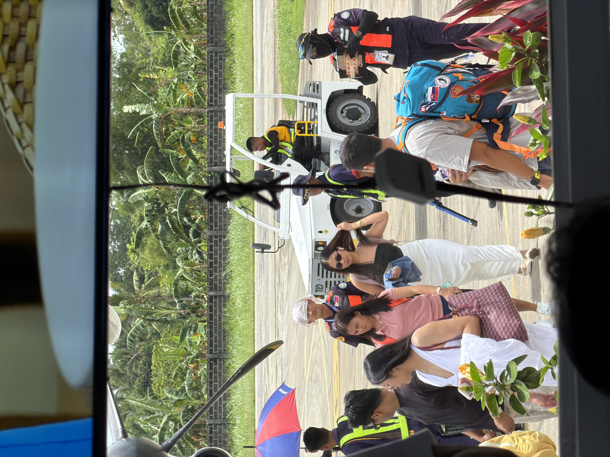 The image shows a group of people disembarking from a small airplane, possibly at Sayak Airport in Del Carmen, Philippines. Passengers are exiting onto the tarmac where a small vehicle is parked. Several ground crew members are present, assisting with luggage. One person holds a colorful umbrella. The environment includes tropical foliage and a paved area with markings, suggesting an airport runway.