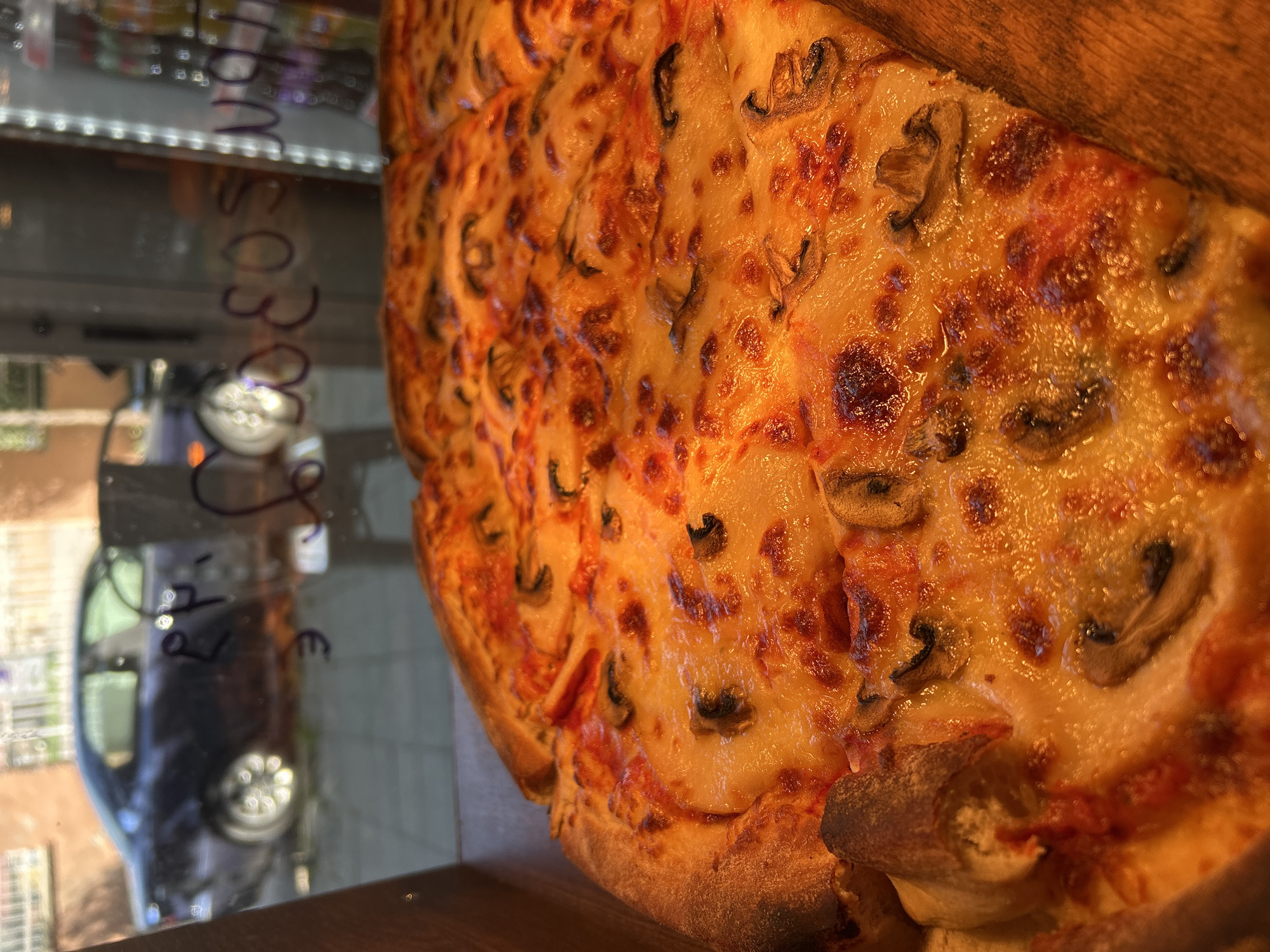 A close-up shot focuses on a mushroom pizza displayed in a wooden box. The pizza is generously topped with melted cheese and sliced mushrooms. Through a glass window behind the pizza, a dark-colored car is visible parked on a street with some buildings in the background. The glass has purple writing on it, which appears to be in Bulgarian. The scene is indoors, likely in a pizzeria or a food establishment.