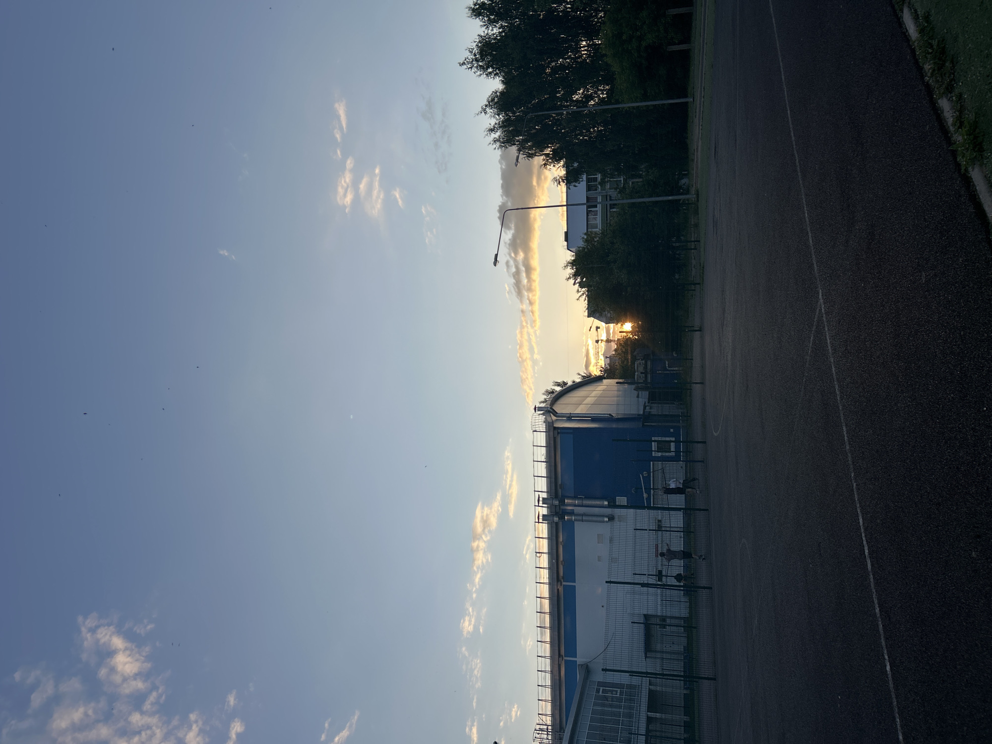 An outdoor sports field with a basketball court is depicted at dusk in Chaikovsky, Russia. The court is made of a dark, textured surface with white boundary lines. A chain-link fence surrounds the court, beyond which a blue and white building with a white roof and railings is visible. To the right of the building, a dense line of tall, dark green trees stands, with a few streetlights scattered amongst them. In the background, an apartment building with a light-colored roof is visible, partially obscured by trees. The sky is a gradient of pale blue to light orange, with wispy clouds, indicating the sun is setting or has just set. Several small dark shapes, possibly birds, are visible in the sky. Three figures, appearing to be individuals, are present on the basketball court, engaged in what seems to be a casual game. One individual is near the basket, while the other two are further away. The overall scene is one of a public recreational space at the end of the day.