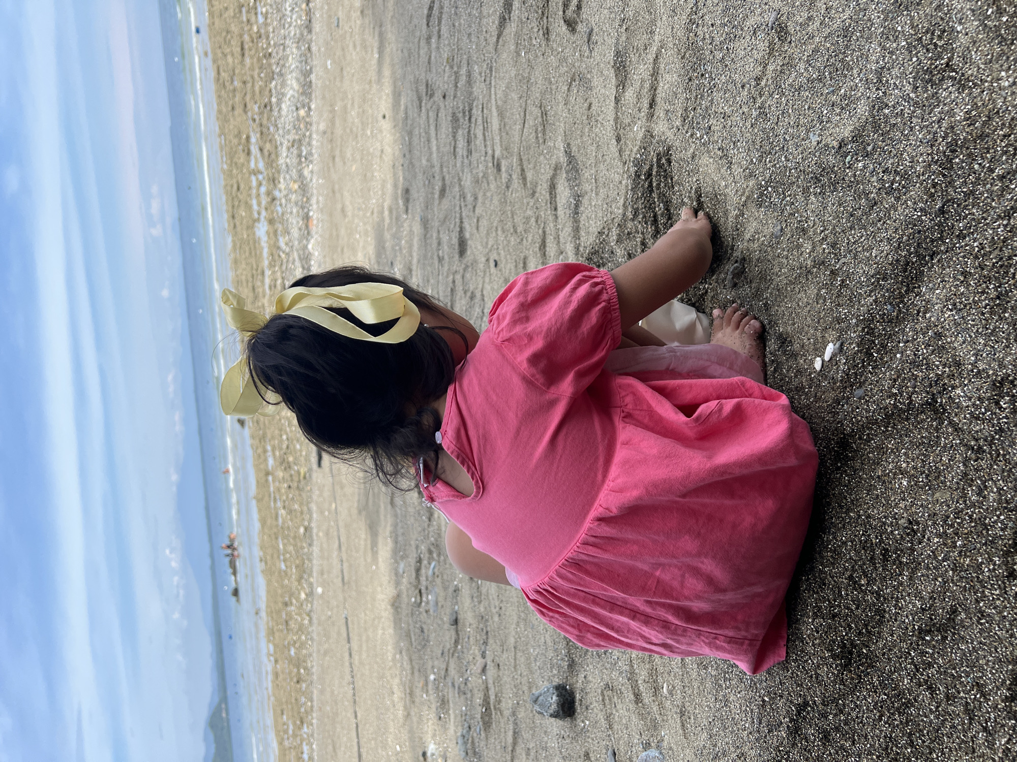 A young girl in a pink dress with a yellow ribbon in her hair is playing on a beach in Banaybanay, Philippines. The girl is sitting on the sand and digging with her hands. The beach is sandy and rocky. The ocean is in the background with people standing in the water. The sky is partly cloudy.