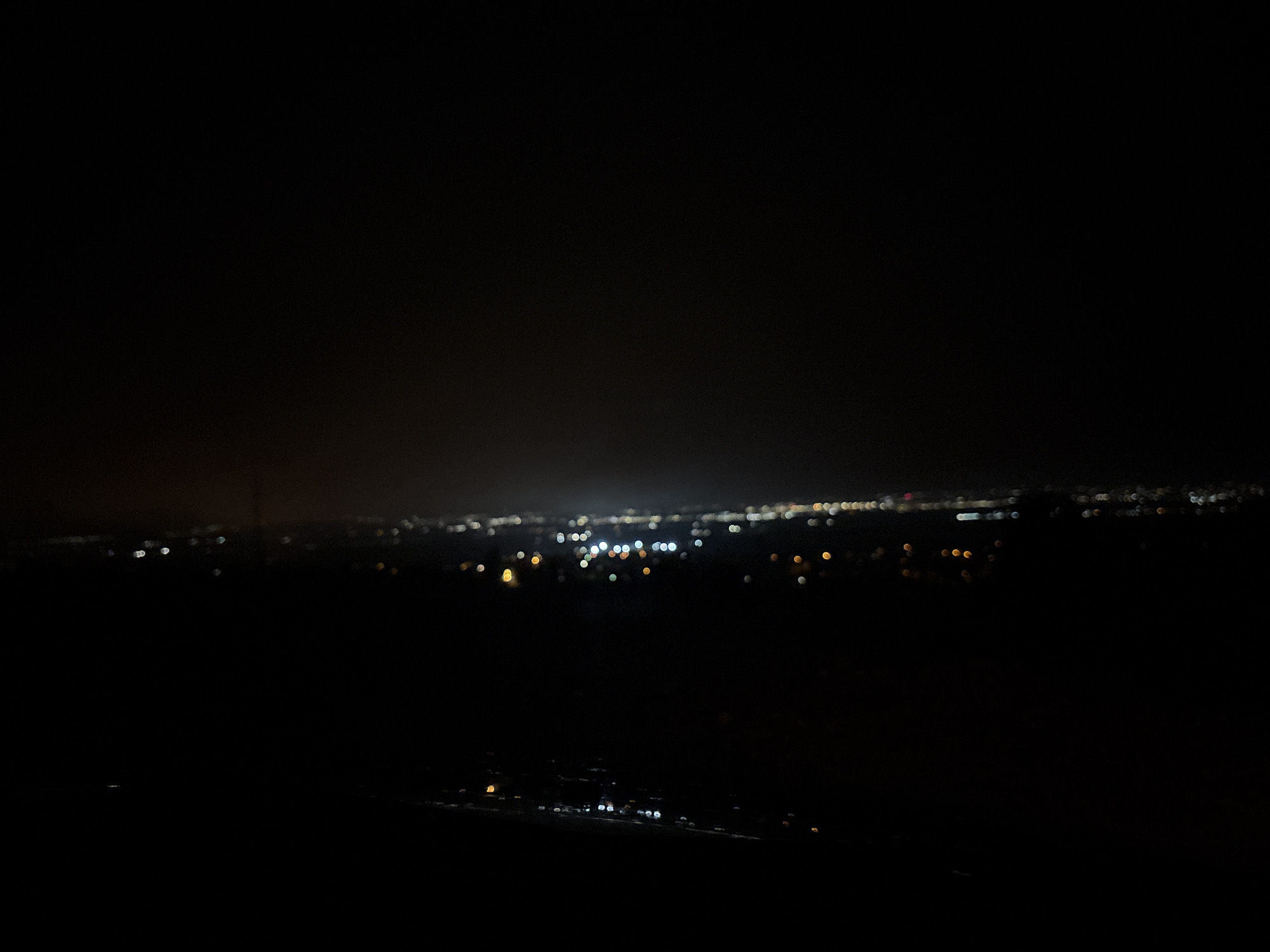 The image presents a dark, expansive night scene, capturing a distant cityscape or town lights from an elevated viewpoint. The upper two-thirds of the photograph are dominated by deep black, representing the night sky, with no stars or other features visible.
Across the mid-ground, a horizontal band of blurred, out-of-focus lights stretches across the frame. These lights appear as various-sized bokeh circles and amorphous blobs, predominantly in shades of white, warm yellow, and orange, with some cooler white points. The distribution of these lights suggests a populated area, possibly a sprawling city or a cluster of towns, rather than a single concentrated urban core, as they are somewhat spread out horizontally. The blur indicates either a shallow depth of field, movement during a long exposure, or simply that the lights are significantly out of focus, enhancing their abstract quality.
The foreground, occupying the bottom third of the image, is almost entirely black and featureless. No specific objects, terrain, or structures are discernible in this area, implying a dark, unlit landscape directly in front of the viewer, such as a hillside or mountain slope.
There are no people, identifiable objects, or visible text in the image. The scene conveys a sense of quiet observation of human activity from a distance, highlighting the artificial illumination against the vast darkness of night. The time of day is clearly night, and there are no discernible weather conditions like rain or fog, though the blur could be partly due to atmospheric haze.