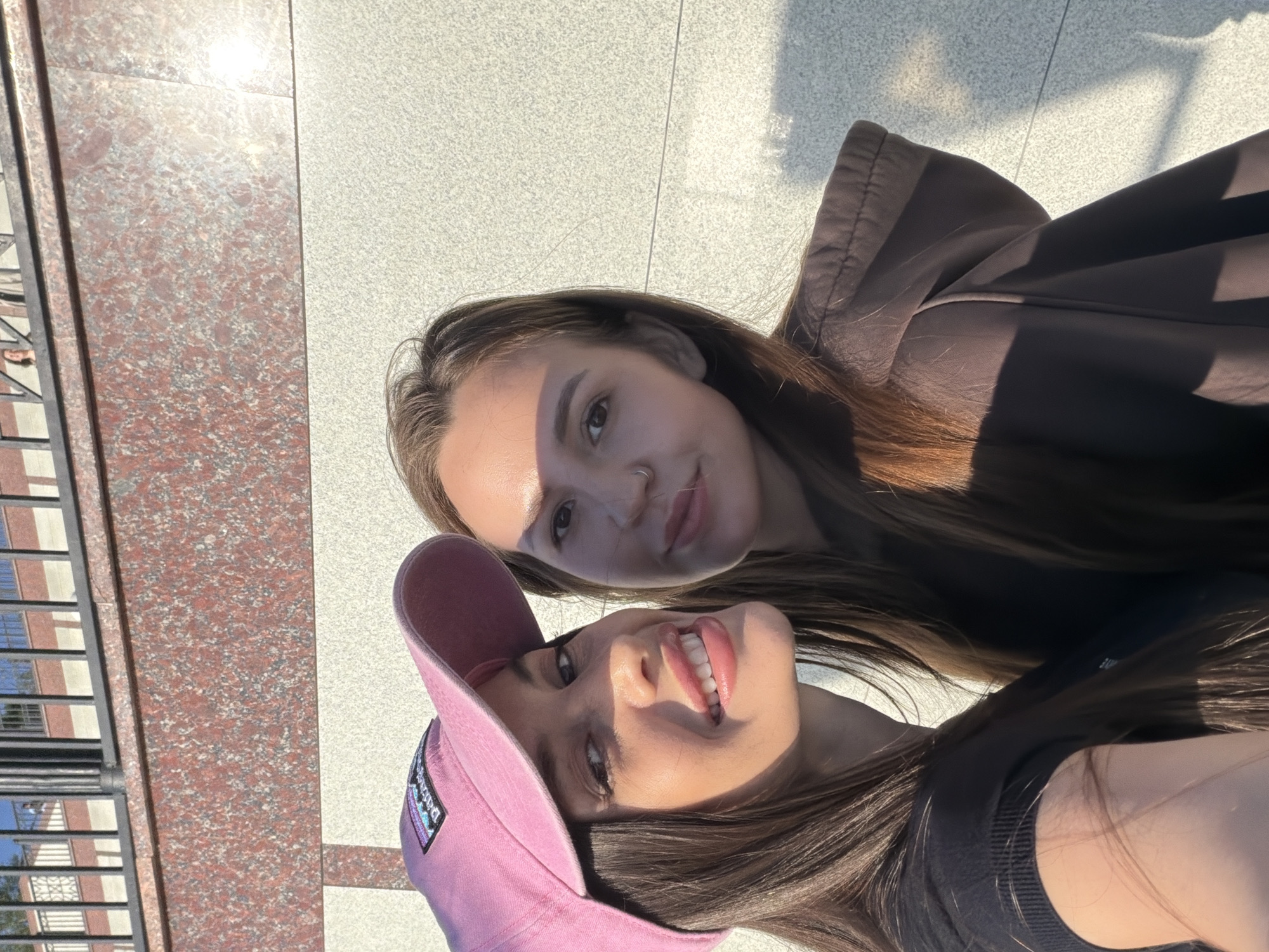 The image, rotated 90 degrees clockwise, features two young women captured in a close-up selfie outdoors on a sunny day in Izhevsk, Russia. The background consists of a two-toned tiled surface: a reddish-brown speckled granite-like section on the left and a lighter, grey speckled section on the right. Strong sunlight illuminates the scene, casting distinct shadows and highlights, particularly on the women's faces.
The woman in the foreground is smiling brightly, showing her teeth, and wears a pink baseball cap with a visible "Patagonia" logo. She has long, dark hair and is wearing a dark top. Slightly behind and above her is the second woman, who has long, dark hair and a nose piercing, offering a subtle smile. She is wearing a brown hooded jacket. Her face is partially shaded by the strong sun.
On the far left, reflections of a multi-story building with numerous windows are visible on a dark, reflective surface, suggesting an urban setting. The overall atmosphere is cheerful and relaxed, depicting a casual moment between the two women.