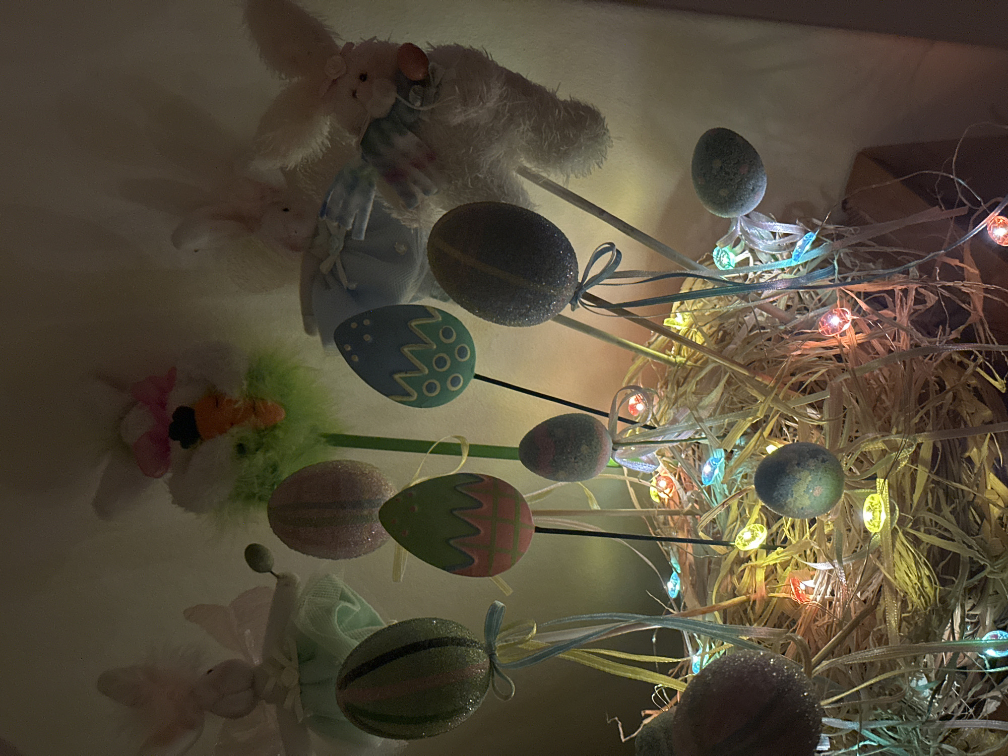 The image shows an Easter-themed display. On the left, there are stuffed bunny decorations, one white and fluffy, and another smaller one. In the center, there are several decorative Easter eggs on sticks, with various designs and colors, some tied with ribbons. On the right, there is a nest-like structure made of straw, illuminated by a string of multi-colored fairy lights. The scene is indoors, illuminated by artificial lighting. There is no indication that this image is taken in Wealden, United Kingdom.