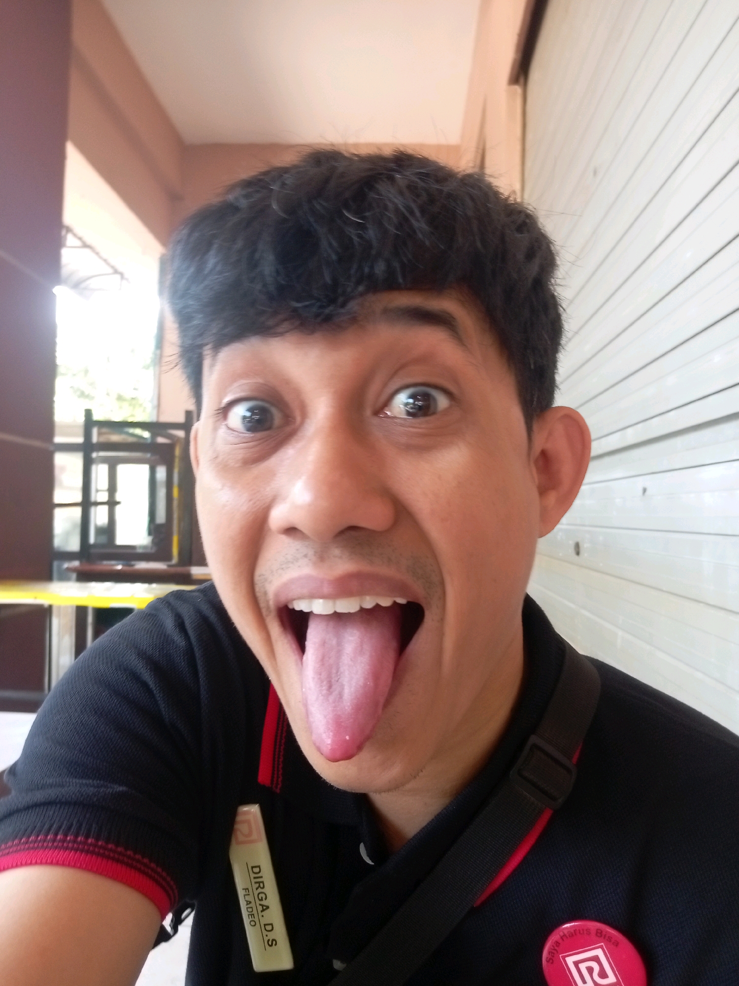 A close-up, low-angle selfie shows a man with dark hair and medium-toned skin sticking his tongue out with his mouth open wide. He is wearing a black polo shirt with red trim on the collar and cuffs. A black strap from a bag or camera crosses his chest. A rectangular name tag is clipped to the left side of his shirt, reading "DIRGA. D.S FLADEO". A round pink pin with a white symbol is on the right side of his shirt. The background consists of a building with pale brown walls and a white rolling door. The overall impression is informal and playful. The name tag suggests the location may be an office or establishment. Based on the name "Dirga" which is common in Indonesia, and the style of the rolling door, the location is likely a public building or commercial space in Batam, Indonesia.