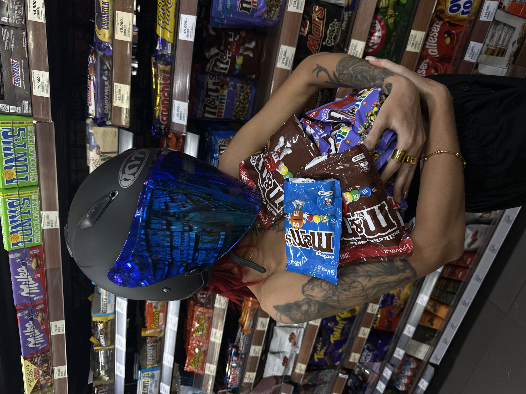 A person with tattoos, red hair, and a helmet is holding multiple bags of M&Ms and Mars bars in a supermarket aisle. The supermarket shelves are stocked with a variety of chocolate bars including Snickers, Dairy Milk, Crunchie, and other confectionery items. Price tags in Indonesian Rupiah are visible on the shelves.
