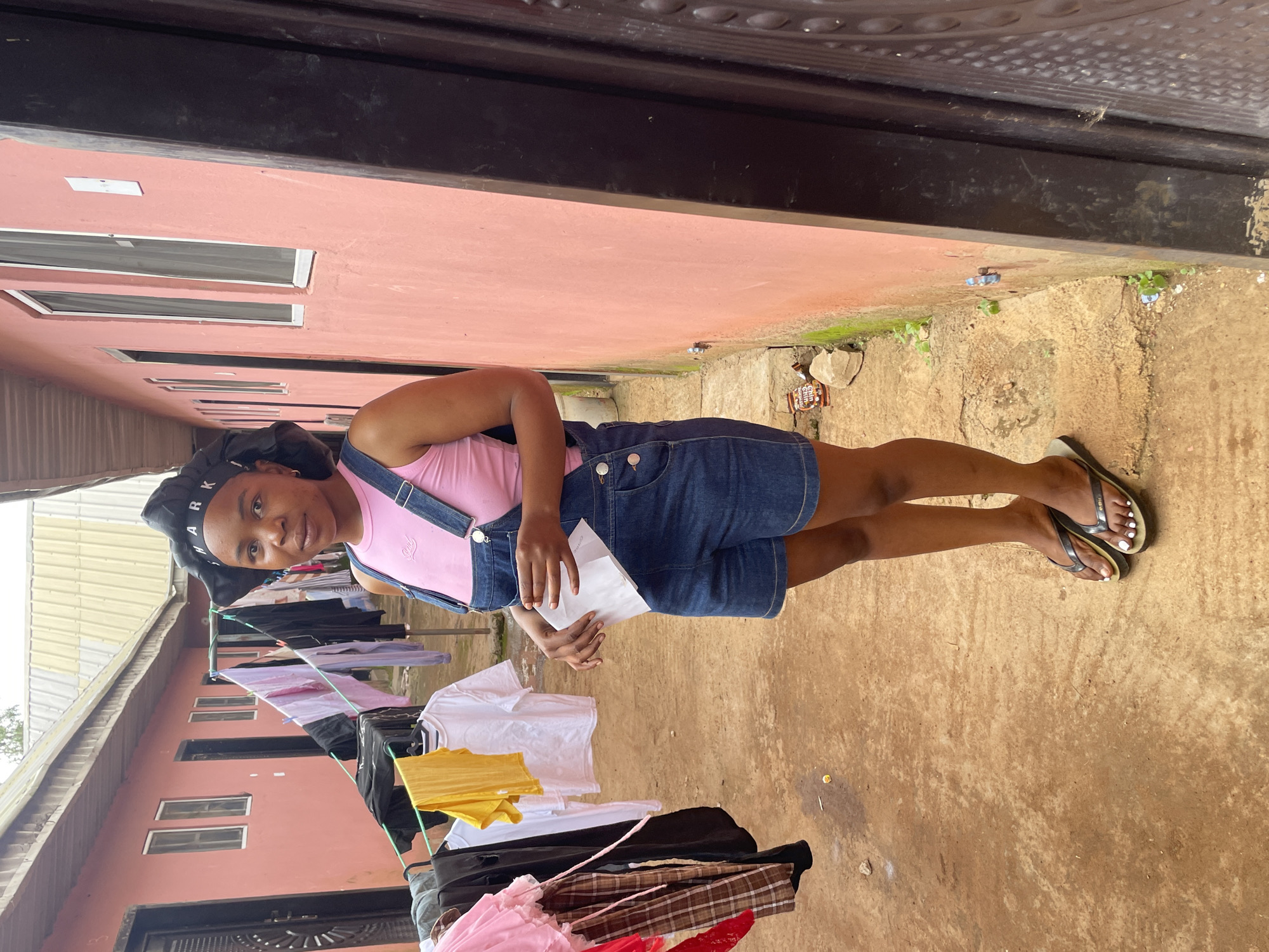 The image depicts a young woman standing outside a building in Durumi, Nigeria. She is wearing a pink tank top and blue denim overalls. Her hair is covered with a black bonnet that has "SHARK" written on it in white letters. She is holding a folded white piece of paper in her hands. To her left, clothes are hanging on a clothesline, with various garments including shirts and pants visible. The building behind her is painted a salmon pink color and has several windows with white frames. The ground is a dirt or gravel surface. In the background, more buildings are visible. The scene appears to be in a residential or courtyard area, with laundry drying and a general outdoor setting.