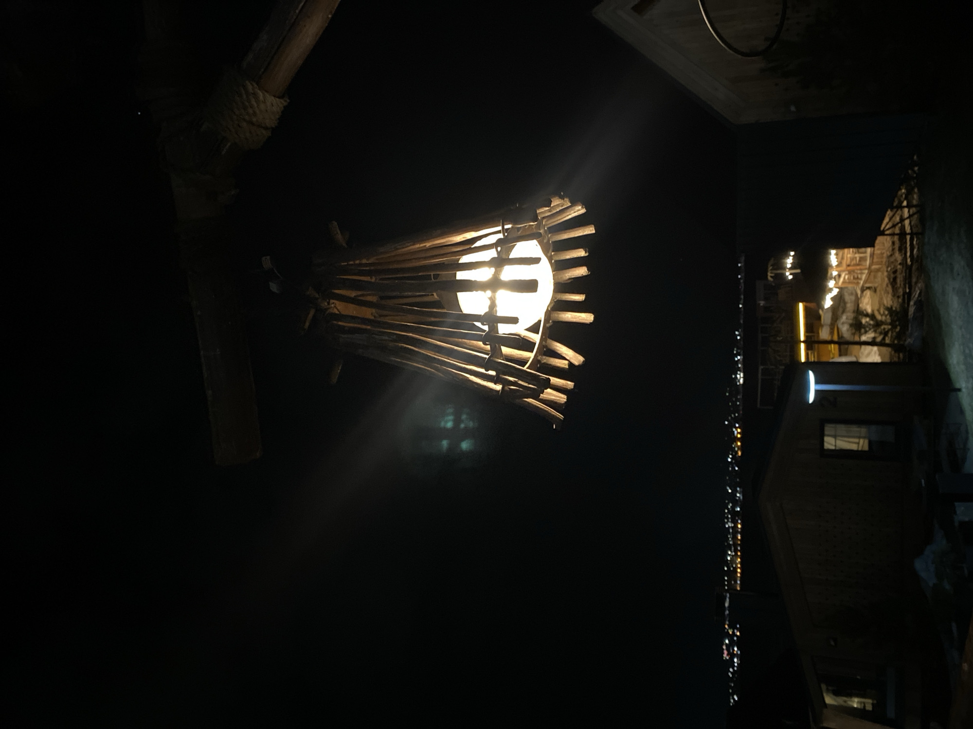 The image shows an outdoor night scene. A hanging light fixture, constructed of wood sticks around a bulb, is brightly lit in the center of the frame. To the right of the fixture, a building is partially visible, with windows and a lighted area near its roof. Further in the background, a line of city lights extends into the distance. Given that Votkinsk, Russia is a small town, there is not a visible match in the image with this specific location.