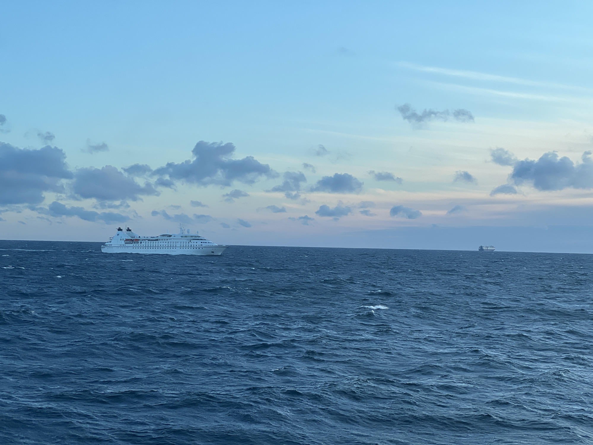 The image depicts a vast expanse of the sea under a twilight sky. The water is a deep blue, with noticeable waves creating a textured surface. In the foreground and mid-ground, the sea dominates the frame. Two ships are visible in the distance. The larger ship, on the left, is a white passenger vessel with multiple decks and visible windows, its hull detailed with orange lifeboats. It is positioned on the left side of the image. Further to the right and much smaller on the horizon is a second ship, also appearing white. The sky transitions from a pale blue near the horizon to a deeper, dusky blue overhead, with scattered clouds, some appearing grey and others with hints of pink and yellow suggesting the fading light of dawn or dusk. The scene is serene and open, with no land features visible. The exact location in the city of Unknown, Poland, is not discernible from the image, as it shows only a seascape.