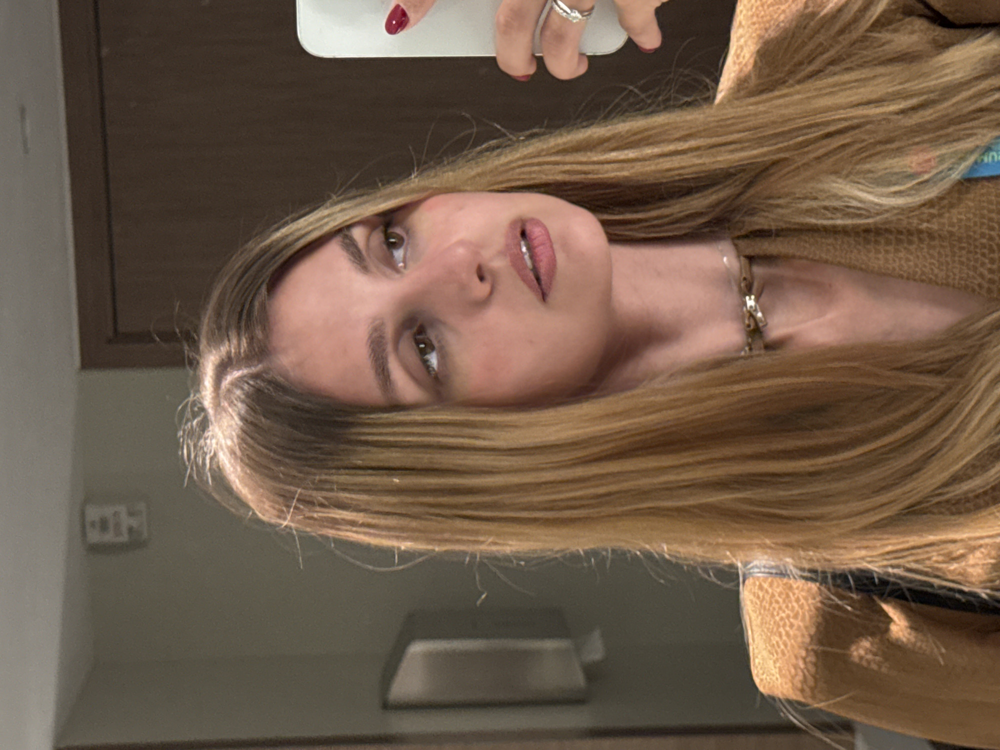 A young woman with long, blonde, straight hair is taking a selfie in a restroom. She is wearing a textured, tan-colored jacket and a choker necklace. Her right hand is visible, holding a white smartphone and wearing a silver ring and a silver necklace. Her fingernails are painted red. The background shows a mirror reflecting the woman and the restroom. A light fixture is visible on the ceiling in the reflection, and a dark wood door is to the right. A silver dispenser is mounted on the wall to the left. The lighting suggests an indoor setting, likely a public restroom.