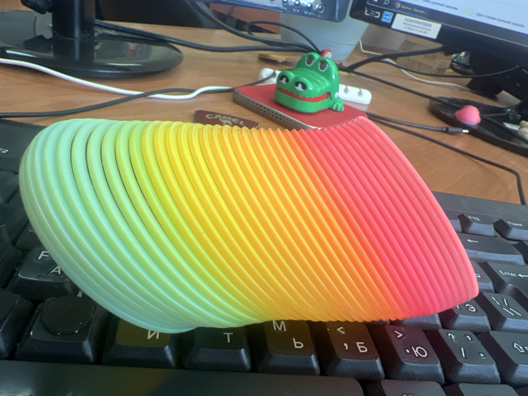 The image depicts a close-up, eye-level view of a rainbow-colored slinky resting on a black computer keyboard. The slinky is positioned horizontally across the keyboard, with its coils transitioning from green on the left to yellow and then to red on the right. A small green toy alligator with large eyes and a red mouth sits to the right of the slinky, on top of a red object, which appears to be a small notebook or notepad. In the background, a computer monitor with some text visible, wires, and a wooden desk surface are present. The scene appears to be an indoor setting, likely a home or office desk. There is no discernible information within the image to place it specifically in Omsk, Russia.