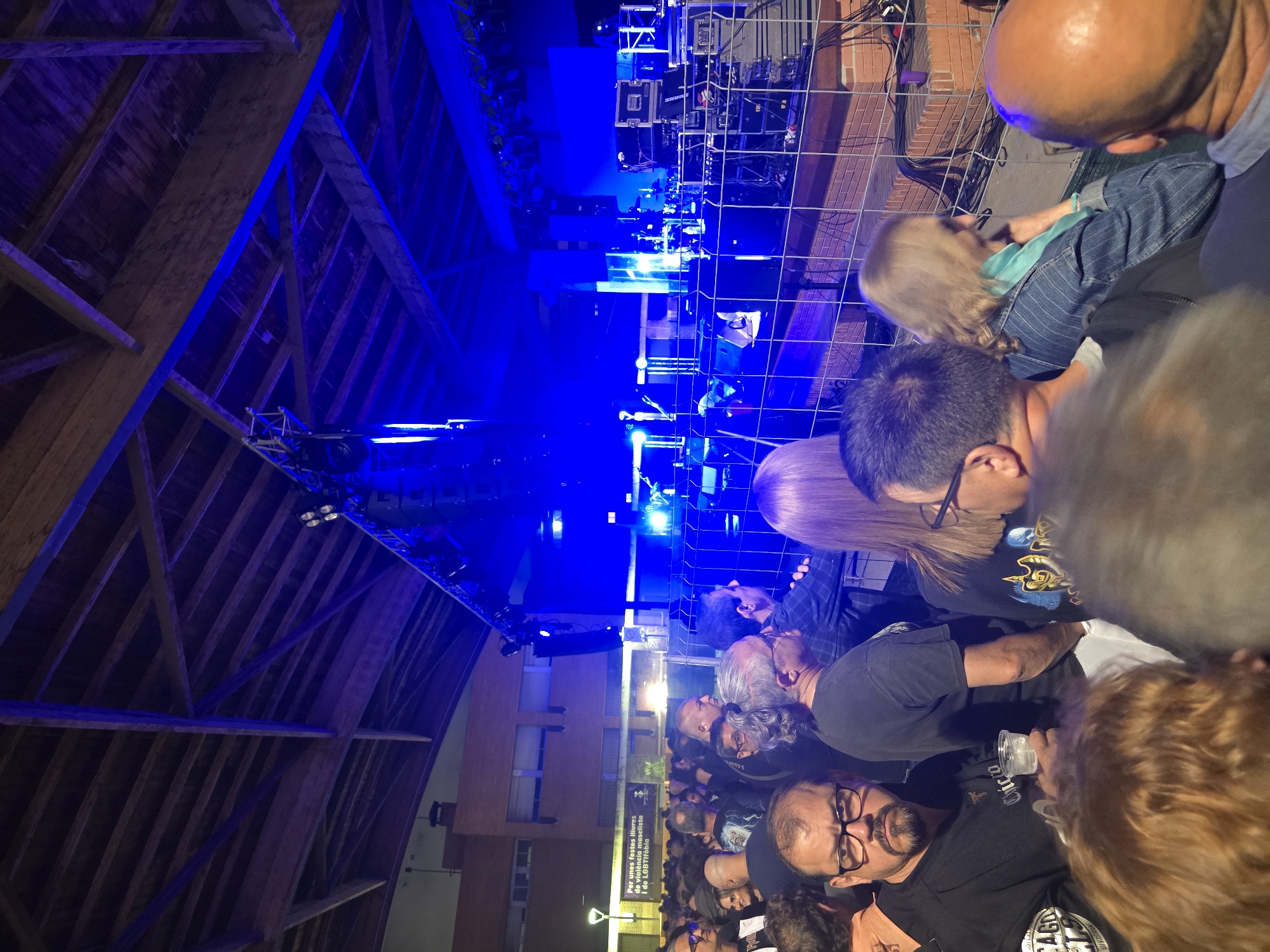 This detailed image depicts an outdoor night concert or live music event in Cornellà de Llobregat, Spain. A crowd of numerous individuals, predominantly adults, is gathered in the foreground and midground, observing a brightly illuminated stage. The stage is bathed in intense blue light, rendering details of the musicians somewhat indistinct, but showing a band performing with a guitarist visible. Stage equipment includes multiple large speakers, lighting rigs suspended from truss structures, a drum kit, and various amplifiers and road cases, all positioned behind a waist-high metal mesh barrier separating the performers from the audience.
Above the stage, a robust wooden ceiling structure with exposed beams and planks is visible, illuminated by the blue light reflecting upwards. In the left background, beyond the crowd and stage area, stands a multi-story brick building featuring multiple rectangular windows. A streetlamp is visible to the left of the building, and a banner is affixed to a wall, displaying the Catalan text: "Per unes festes lliures de violència masclista i de LGBTI-fòbia" (For parties free of sexist violence and LGBTI-phobia).
In the immediate foreground, several audience members are visible. One prominent individual, a man with glasses and a dark beard, wearing a black shirt with "CHICAGO" text partially visible, holds a clear plastic cup containing a light-colored beverage. Another man with gray hair in a ponytail is visible behind him. Towards the right, a woman with blonde hair, wearing a denim jacket and a light blue scarf, observes the stage. The attendees are standing, generally facing the stage, indicating their engagement with the performance. The overall scene suggests a vibrant public gathering for live entertainment.