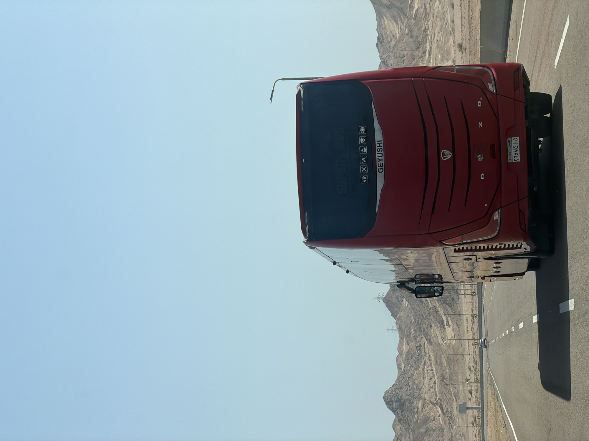 The image shows the rear of a red Geyushi bus on a highway in Sharm El Sheikh, Egypt. The bus is the primary subject, dominating the foreground and center of the frame. Above the rear windshield, text reads "SUPER BUS" and icons for VIP, WC, Wi-Fi, and a power outlet are visible. The bus's license plate is in Arabic script, indicating "Egypt." The road stretches ahead into a desert landscape with rugged, barren mountains in the background. A single white car is visible in the distance on the same highway. The sky is clear and blue. The scene appears to be during daylight.