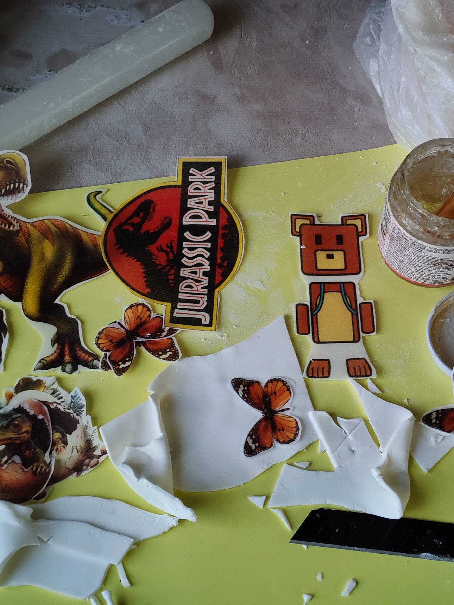 The image depicts a crafting or decorating scene, likely related to cake decorating, situated on a pale yellow surface. Visible are various cut-out images and decorative elements. These include: a dinosaur, the "Jurassic Park" logo, butterfly images, a pixelated bear, and a dinosaur hatching from an egg. There are also fragments of white material, possibly fondant or sugar paste, scattered around. A jar of clear gel, possibly for glazing or adhering decorative elements, and a small black blade are also present. A rolling pin is visible in the upper left of the frame. The setting appears to be a work surface or table. The location is Unknown, Unknown.