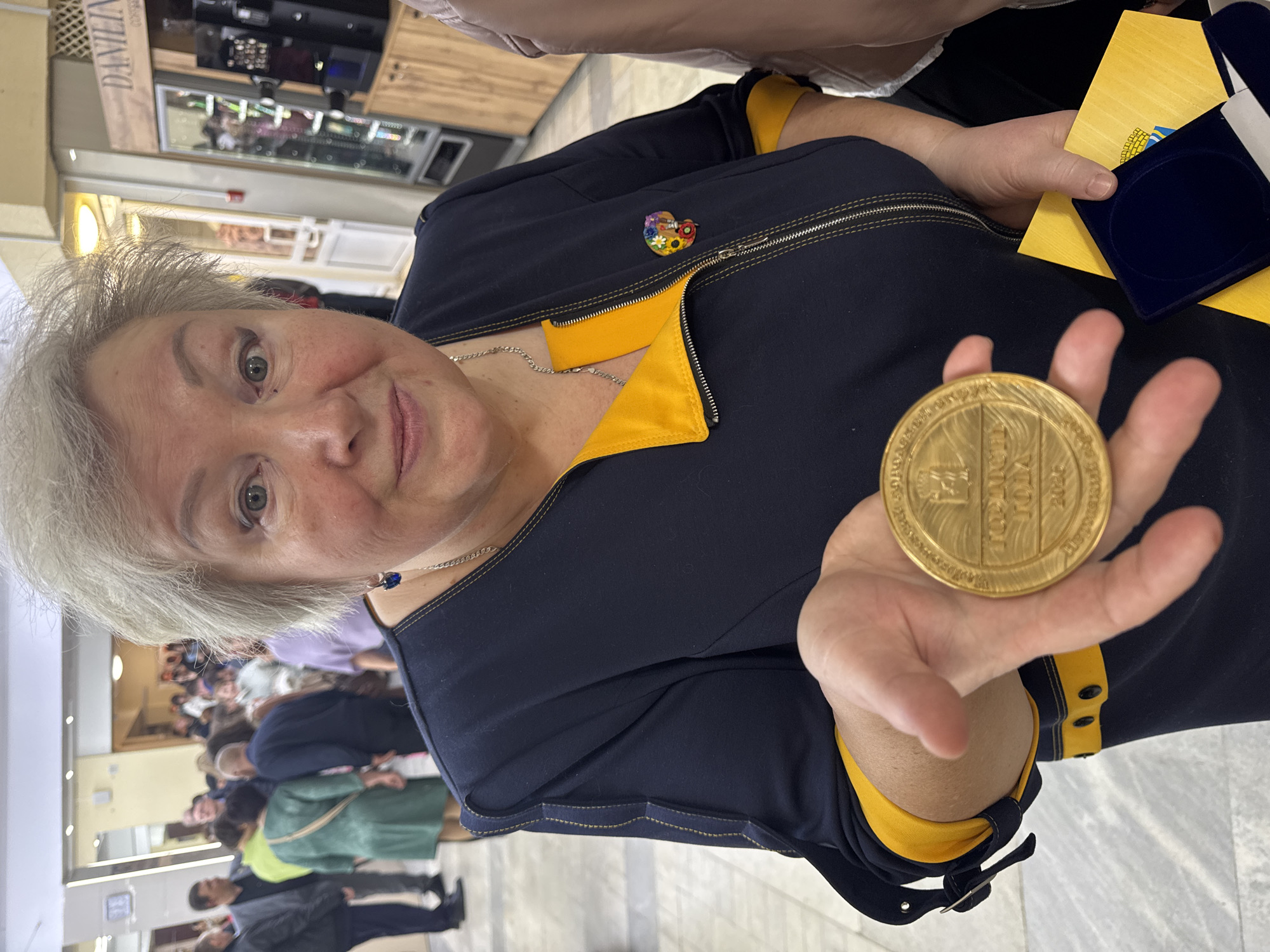 The photograph shows a mature woman holding a gold medal in her outstretched palm. The medal appears to be a commemorative one. Behind her is a crowd of people and what appears to be an indoor public space, such as a mall. The woman has short, gray hair and is wearing a dark-colored jacket with yellow trim, adorned with a colorful pin. She is also wearing a necklace with a blue bead. In her other hand, she holds a folded yellow paper and a blue velvet box. The image suggests a formal event or award ceremony. According to the inscription on the medal, the event is taking place in the city of Chaikovsky, Russia.