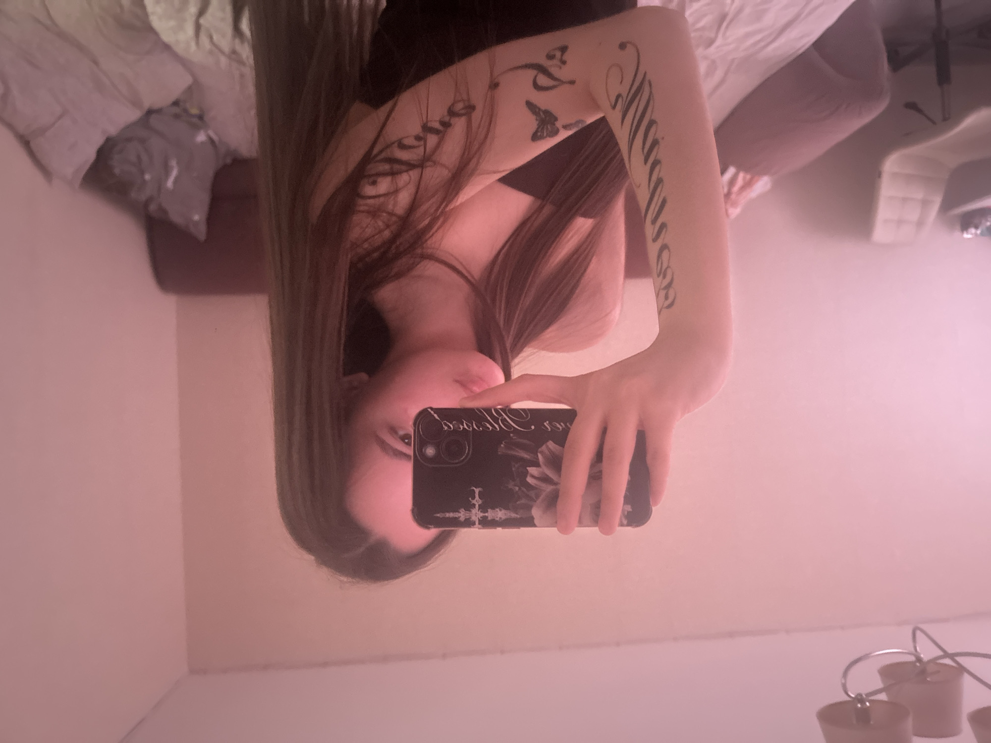 A selfie-style image depicts a woman in an indoor setting, likely a bathroom. The image is presented upside-down. The woman is holding a cell phone with a floral-patterned case. Part of her face is visible, including an eye and part of her nose and mouth. She has long brown hair. A tattoo is visible on her arm, featuring script and a butterfly. In the background, a bed with bedding is partially visible, along with a white chair or stool. The environment appears to be a bathroom based on the presence of light fixtures in the lower portion of the frame. There is no data about the location in the city of Chaikovsky, Russia.