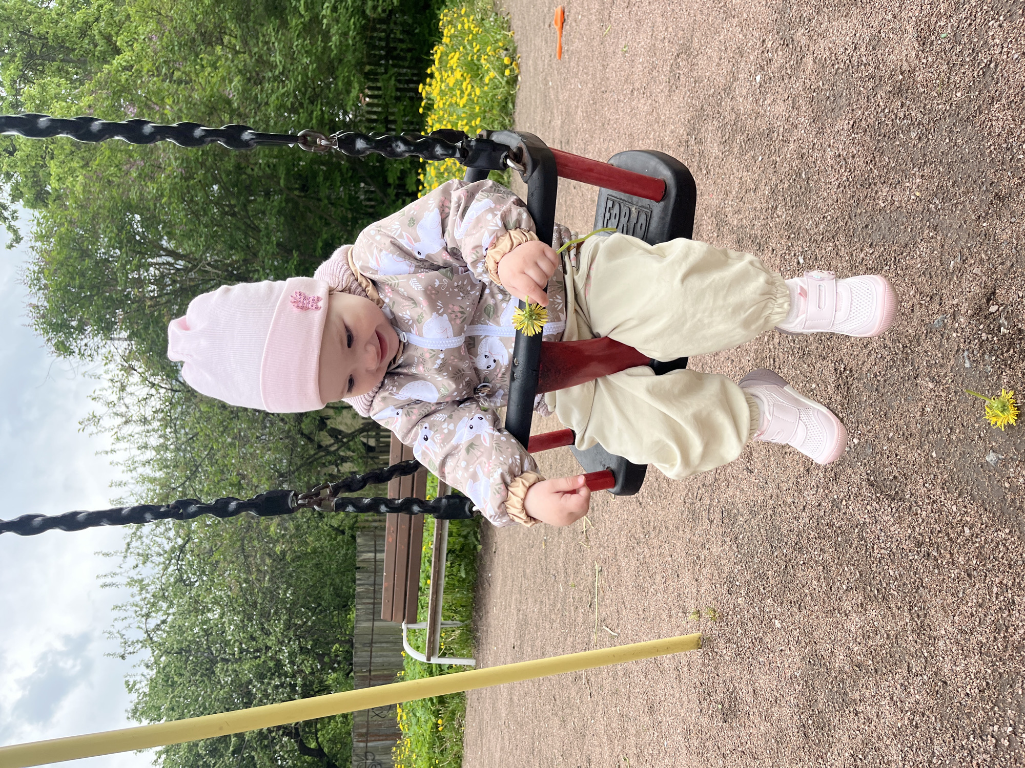 A baby is on a swing in a playground setting. The baby wears a pink hat, a patterned jacket with white bunny designs, beige pants, and pink sneakers. The baby is holding a dandelion. The swing has a red and black frame and black chains. The ground is gravel. In the background, there are trees, yellow flowers, a wooden fence, and a park bench. This image was taken in the city of Pushkin, Russia.