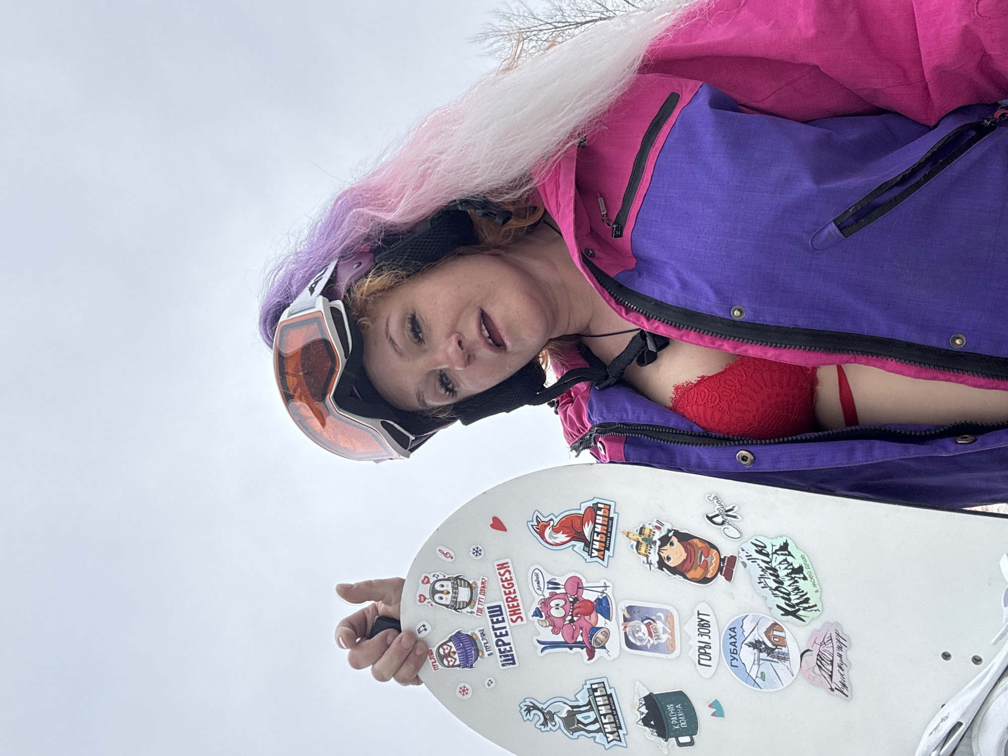 A person, likely female, is featured in a low-angle photograph, appearing to look down at the viewer from above, with the image itself being rotated counter-clockwise. The subject is dressed in winter sports attire, including a vibrant pink and purple snowboard jacket that is partially unzipped, revealing a red lace bra underneath. A ski helmet with orange-tinted goggles pushed up onto its brim covers their head, and long, multi-colored hair (white, pink, and purple) flows out from behind.
The person holds a white snowboard, which is extensively covered with various stickers. Many of these stickers name prominent Russian ski resorts and mountain regions, including "ШЕРЕГЕШ" (SHEREGESH), "ХИБИНЫ" (Khibiny), "Домбай" (Dombay), "КРАСНАЯ ПОЛЯНА" (Krasnaya Polyana), "АРХЫЗ" (Arkhyz), and "ГУБАХА" (Gubakha). Other texts on the stickers include "ГОРЫ ЗОВУТ" (Mountains call), "В ПУХЛЯКЕ" (In powder), "ГДЕ ТУТ ДВИЖ?" (Where's the party?), and "ПУХЛЯШ" (Fluffy/Powder), along with graphical elements like penguins, a fox, and a monster on skis. One sticker reads "Курдискумчорр в моей душе" (Kurdiskumchorr in my soul).
The background is a bright, overcast sky, with faint bare tree branches visible, suggesting an outdoor, cold-weather setting typical of a ski or snowboard resort during the daytime. The person’s expression is neutral, with a direct gaze towards the camera. The ensemble of snowboarding gear and the specific resort names on the stickers indicate an activity related to winter sports, possibly at Sheregesh or another mountain destination in Russia, like those found near Chaikovsky.