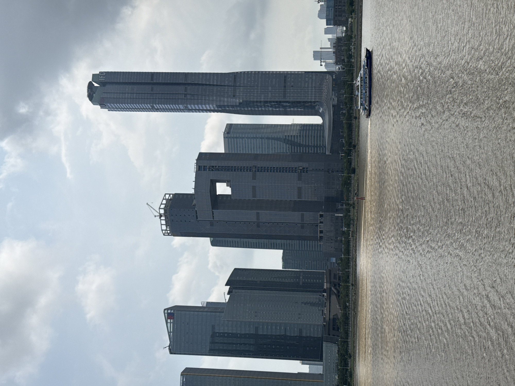 The image shows a skyline of modern buildings next to a body of water. The buildings are a mix of skyscrapers and other high-rise structures, with construction visible on at least one of them. A boat is present on the water. The sky is overcast.
