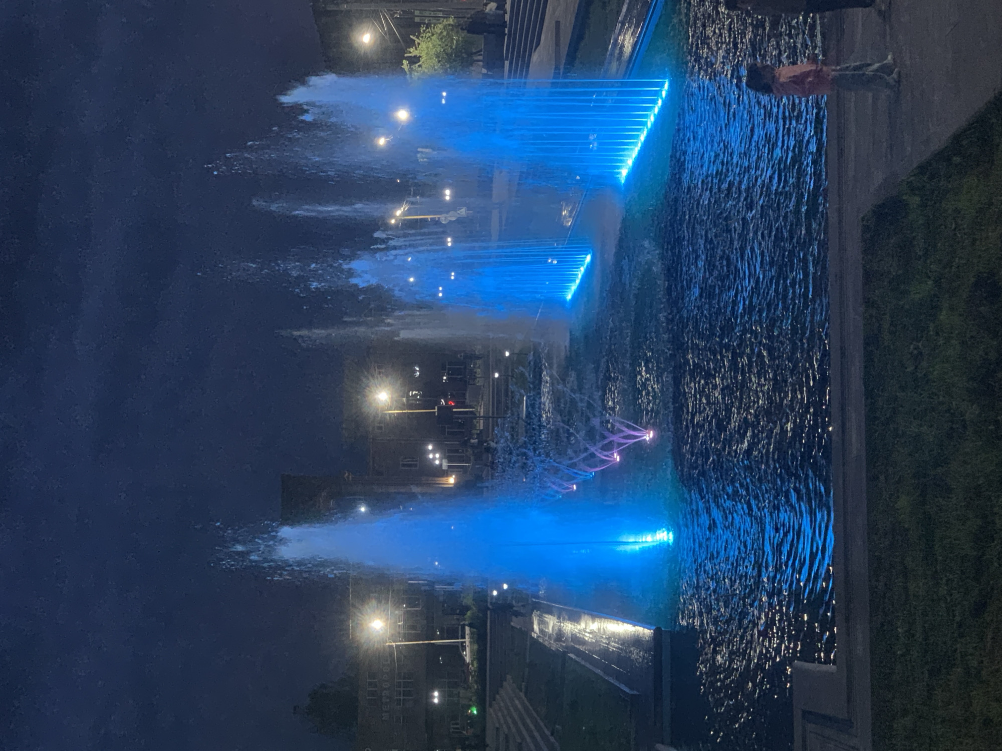 This nighttime image captures the Gyumri Cascade in Gyumri, Armenia, a terraced complex featuring multiple illuminated water fountains. The sky is dark blue, indicative of late evening or night.
In the center-left, a prominent, conical fountain sprays a high, wide column of water intensely illuminated in bright blue. To its immediate right, a series of smaller, lower water jets are lit with vibrant pink and purple lights. Further to the right and closer to the foreground, two distinct rectangular fountain structures project numerous vertical jets of water, all glowing with vivid blue light. The water in the fountain basins reflects these blue hues and shows visible ripples.
The background features dimly lit urban architecture. On the left, a multi-story building with the word "METRO" partially visible on its facade is illuminated by a bright, warm-toned streetlight. Other dark buildings line the background on both sides. Multiple streetlights are scattered throughout the scene, providing localized illumination to pathways and background areas.
On the lower right, two figures stand on a paved pathway observing the fountains. The most discernible figure is a child wearing a light pink jacket and light blue or gray pants, positioned with their back partially to the camera. Adjacent to the child, a taller, indistinct figure in dark clothing is partially visible. Further back, near the buildings and steps, several other dark, indistinct figures are present, suggesting light pedestrian activity.
Stone or concrete steps and pathways ascend on both the left and right sides of the fountain complex. Patches of dark green grass are visible in the immediate foreground and along the edges of the pathways. A small tree with dark green foliage is situated on the right side of the background, near a set of steps.