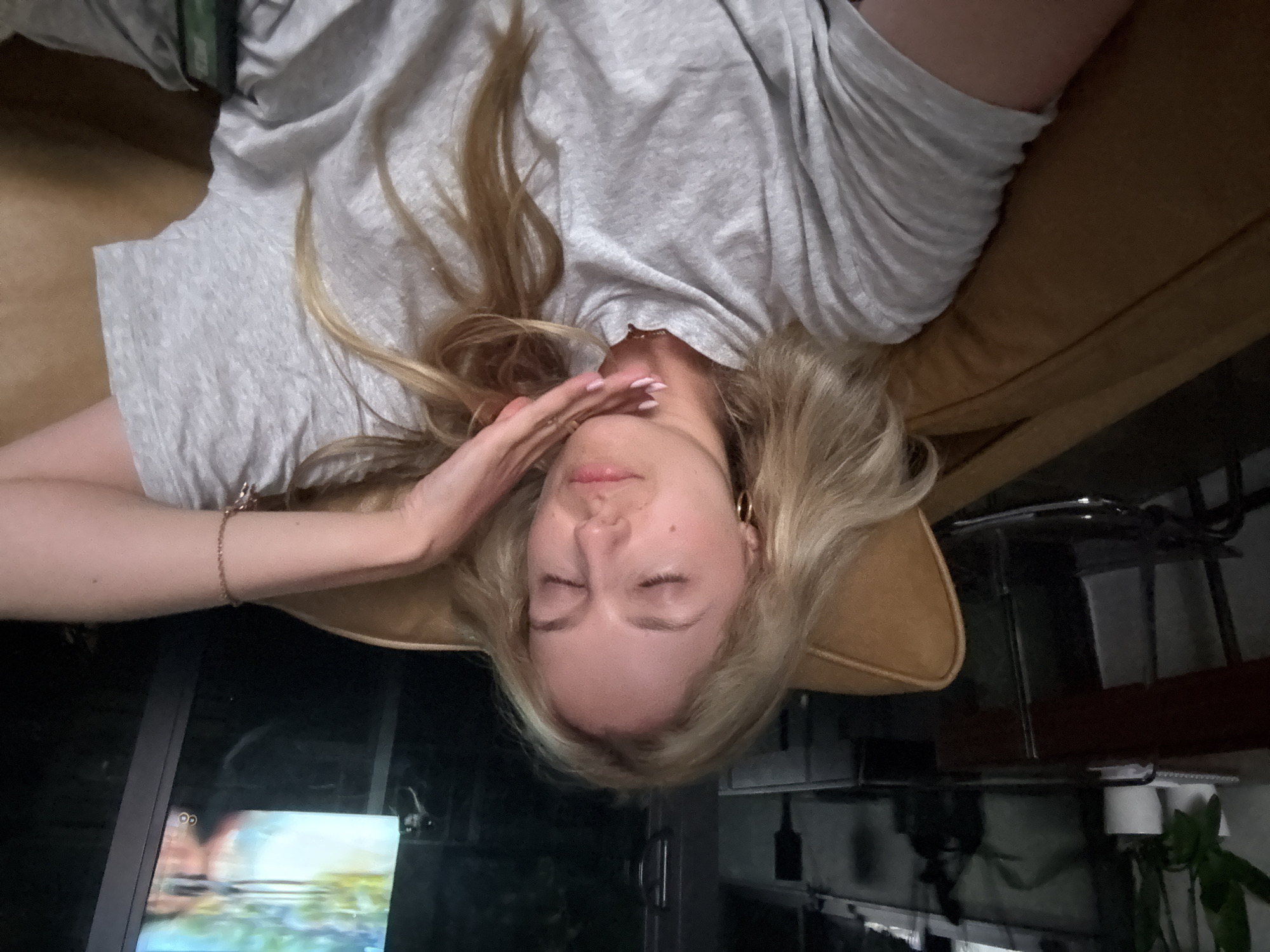 In Miami, Florida, United States of America, a blonde woman is reclining on a tan couch, her eyes closed and her head tilted back. She is wearing a grey t-shirt and her right arm is bent, with her hand resting on her chin, displaying manicured fingernails. A thin bracelet adorns her wrist. The background features a television screen displaying a vibrant, colorful image, and dark interior elements. To the left, a dark wooden shelf holds two potted plants in white containers, suggesting an indoor domestic setting.