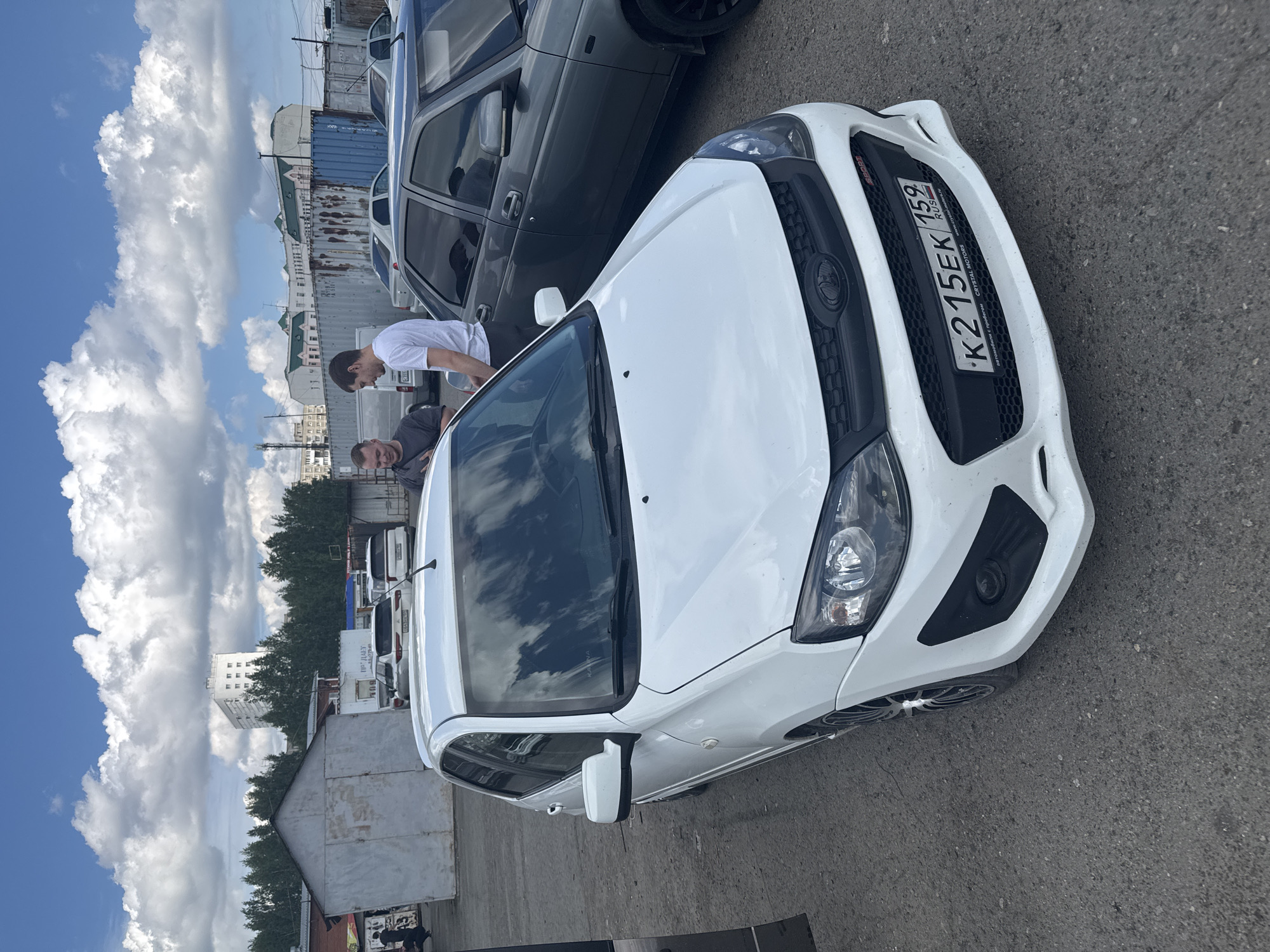 A medium shot captures a white Lada Granta, registered in the Perm region of Russia with the license plate "K 215 EK 159". The car is parked on an asphalt surface, with a gray sedan parked adjacent. Two men are visible near the cars. The background reveals a cityscape with buildings and partly cloudy sky.