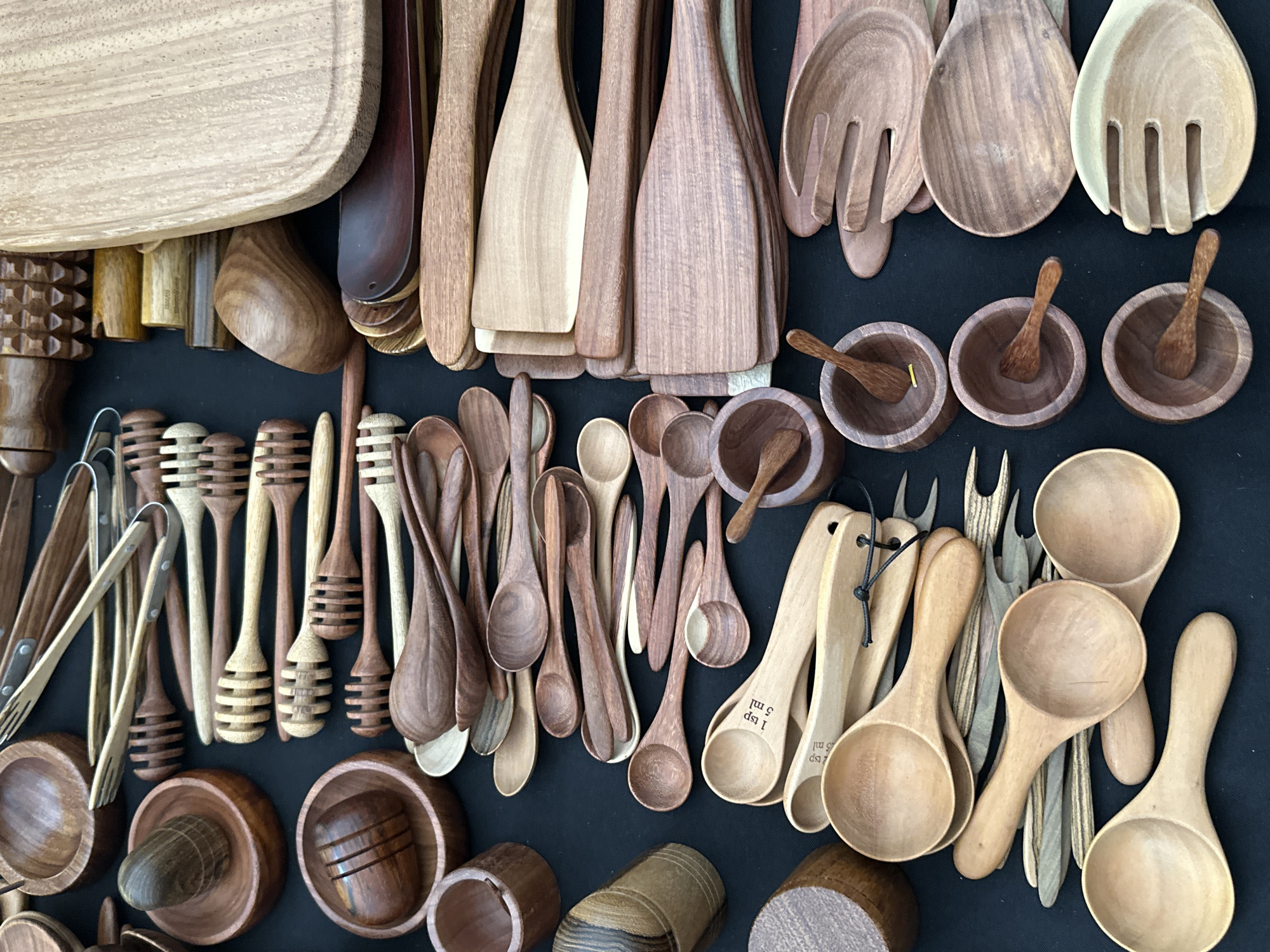 The image displays a flat lay arrangement of numerous wooden kitchenware and home decor items on a black, matte surface. The scene is set in Mexico City, Mexico.
The composition features a variety of wood tones, ranging from light beige to deep reddish-brown and dark chocolate brown, showcasing different wood grains and finishes.
**Key objects observed include:**
* **Top Left:** A dark brown wooden bowl containing a rounded wooden masher or pestle, and a set of light wooden tongs with silver metal hinges.
* **Upper Center Left:** Several light-colored wooden honey dippers, characterized by their ribbed, cylindrical ends, are neatly arranged in rows. Above them, more wooden tongs with metal hinges are visible.
* **Mid-Left:** A large, dark brown wooden mortar holds a smooth, dark wooden pestle. Another dark brown wooden bowl contains a small, ribbed wooden object. Several small, cylindrical wooden containers or bases are also present.
* **Center:** A prominent collection of smooth wooden spoons and spatulas of varying sizes and wood types, some stacked, others individual. A light-colored wooden measuring spoon clearly displays "1 tsp 5 ml" inscription. Interspersed are small, multi-pronged wooden fork-like utensils.
* **Lower Center:** Several small, dark brown wooden bowls, each containing a miniature wooden spoon, are clustered together.
* **Top Right:** A large, light-colored wooden cutting board with a rounded edge and groove along its perimeter dominates the upper right quadrant.
* **Mid-Right:** Stacks of flat wooden spatulas and turners, some in a lighter wood and others in a darker, reddish-brown wood, are neatly arranged.
* **Lower Right:** Two sets of large wooden salad serving utensils (a spoon and a multi-pronged fork), one in a dark brown wood and the other in a lighter wood, are visible towards the bottom right.
All items appear new and unused, likely displayed for sale in a market or retail setting. No people are visible.