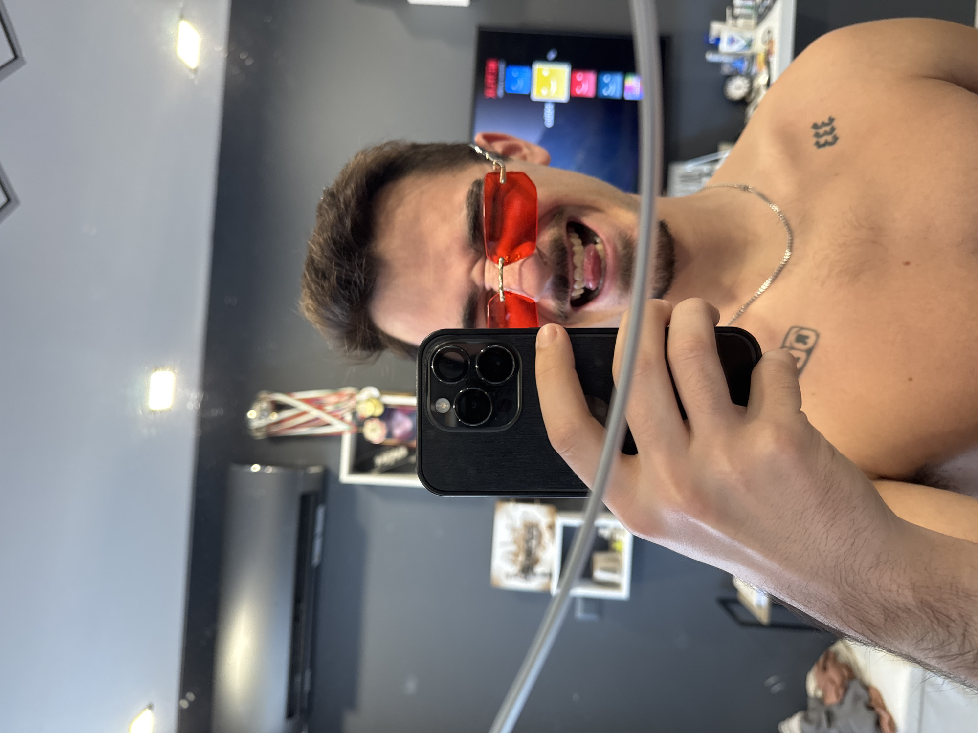 A shirtless man with tattoos, a silver chain, and red sunglasses is taking a mirror selfie indoors. He is holding a black smartphone. The reflection shows a television, artwork, and other objects. The location cannot be determined from the image.