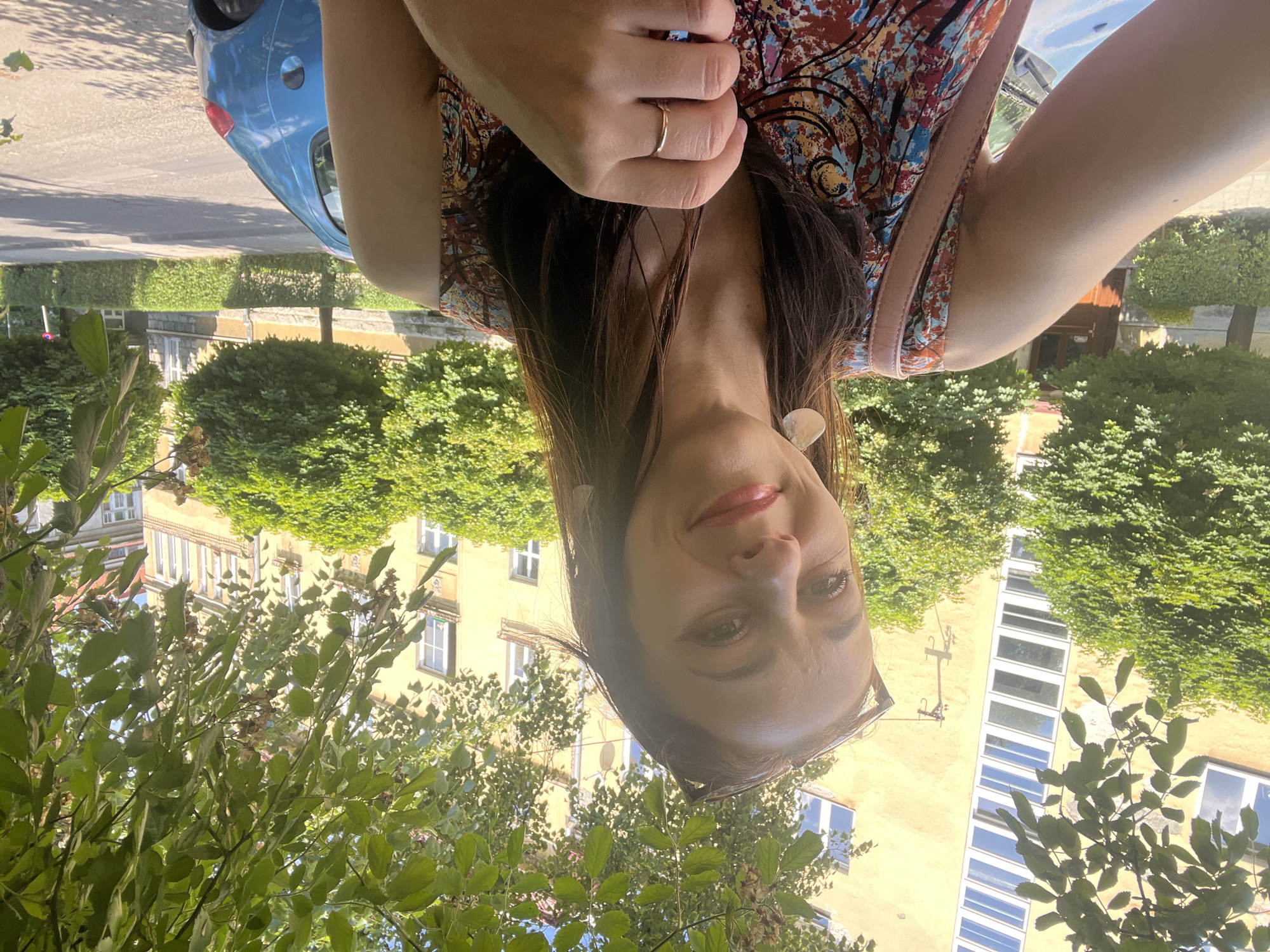 The media file depicts a woman in a close-up shot, with her face positioned near the bottom of the frame. She appears to be outdoors, with lush green foliage in the foreground and a yellow building with multiple windows in the background. A blue car is visible on the left side of the frame, parked on a street. The woman is wearing a colorful patterned dress and a delicate gold ring on her finger. Her expression is a gentle smile, and her eyes are looking towards the camera. The lighting suggests it is daytime, possibly late afternoon, given the warm tones. The overall scene conveys a sense of casual enjoyment and relaxation. There is no visible text in the media file.