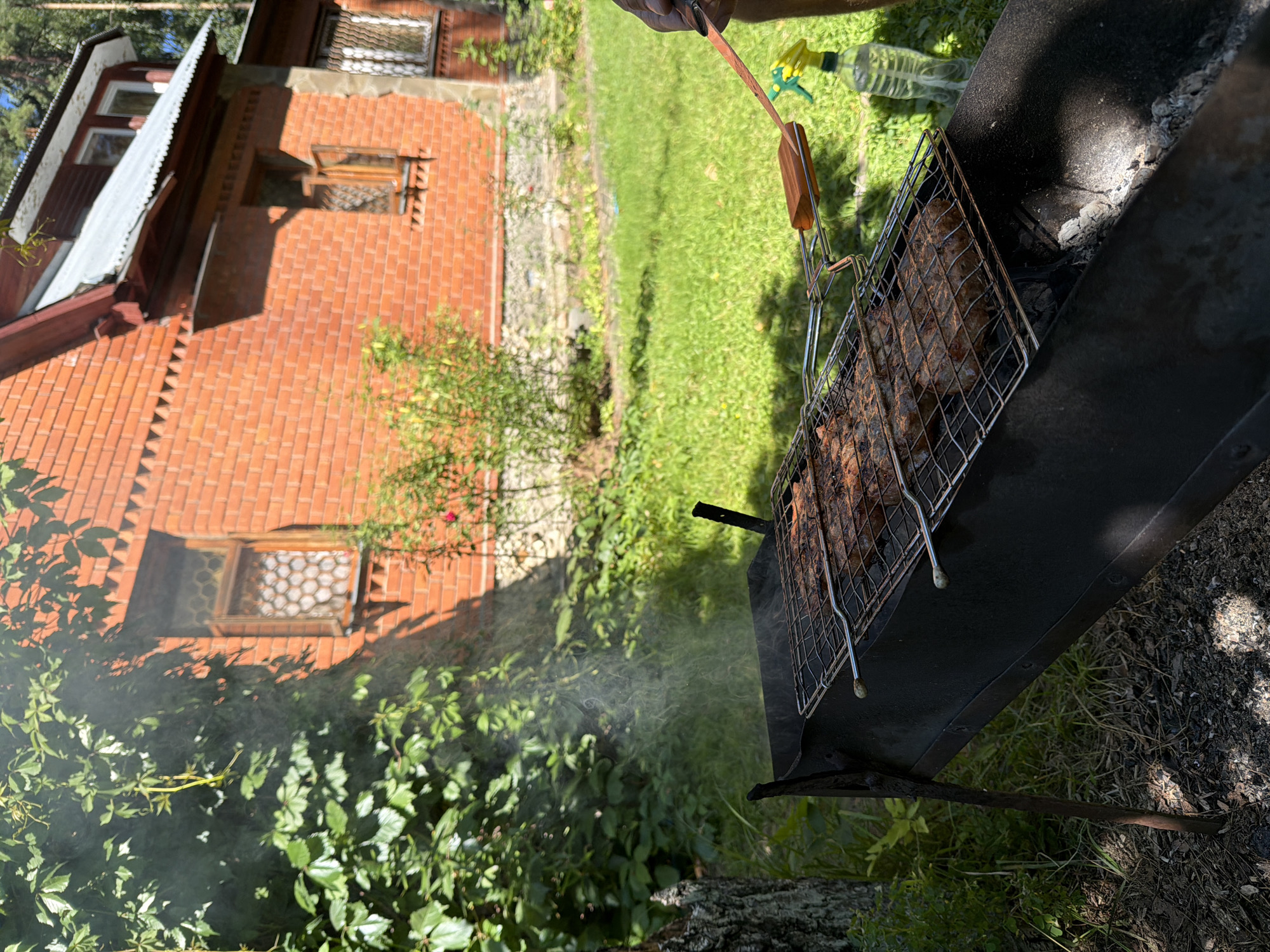 In the city of Udelnaya, Russia, a barbecue is in progress in the backyard of a red brick house. The house, with its wooden window frames and corrugated metal roof, stands in the background, partially obscured by lush green foliage. In the foreground, a metal grill is filled with meat being cooked over hot coals. Smoke rises from the grill, creating a hazy effect. A pair of barbecue tongs and a spray bottle are placed on the grill. The scene is set on a sunny day, with shadows cast on the ground, indicating a warm, outdoor activity.