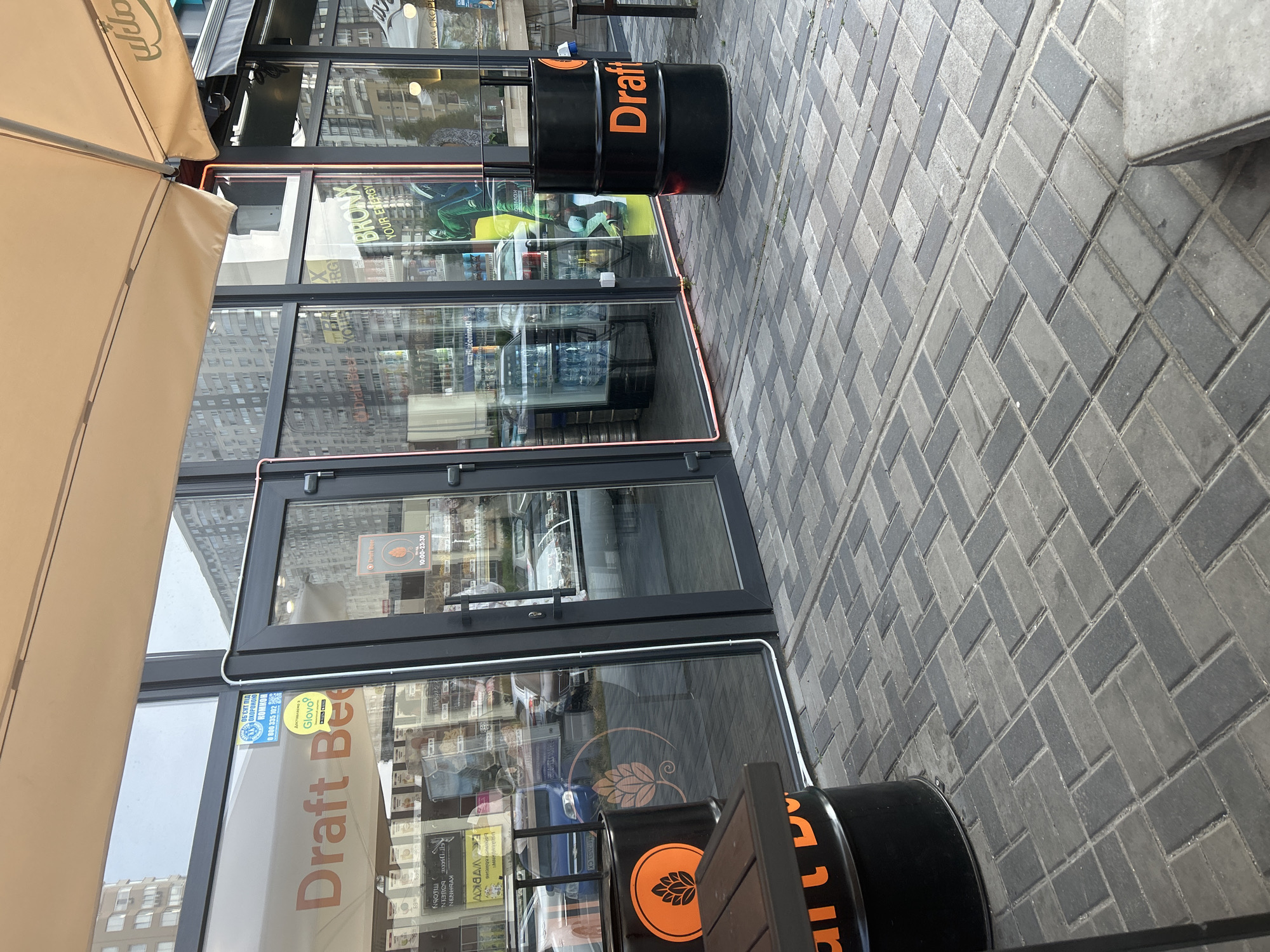 The image captures the exterior of a "Draft Beer" establishment in Brovary, Ukraine. The building features large glass windows showcasing merchandise and refrigeration units inside. Several black barrels, adorned with orange logos of a hop cone and the text "Draft Beer," are placed outside as decorative elements. One barrel is positioned near a dark wooden table. The entrance consists of double glass doors with dark frames, one of which is slightly ajar. A sign on the glass displays operating hours from 10:00 to 23:30. The ground in front of the establishment is paved with interlocking gray and dark gray bricks laid in a herringbone pattern. An awning, partially visible at the top, provides shade. Reflections in the glass indicate a building facade and sky in the background. The overall scene depicts a commercial establishment on a street.