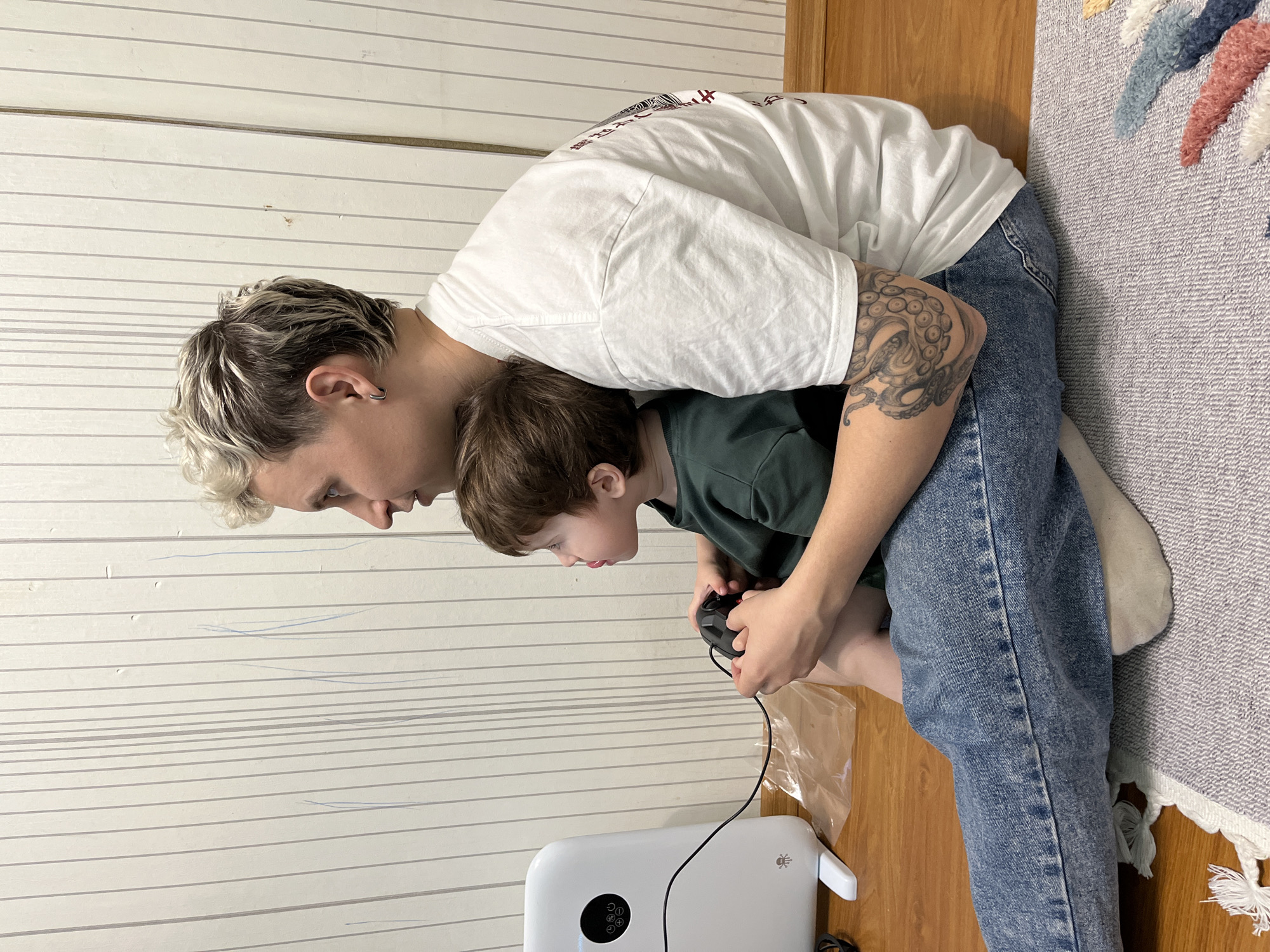The image depicts a light-skinned adult with short, dyed hair and a visible tattoo on their arm, wearing a white t-shirt and blue jeans. They are leaning over a light-skinned child in a green shirt, who is holding a black video game controller. A white, rounded electronic device with a black circular display is visible on the floor near the bottom of the image, with a black wire extending from it towards the controller. The background consists of a wall with horizontal, light-colored paneling and a section of a colorful rug on a wooden floor. The location, Chaikovsky, Russia, cannot be forensically confirmed from this image.