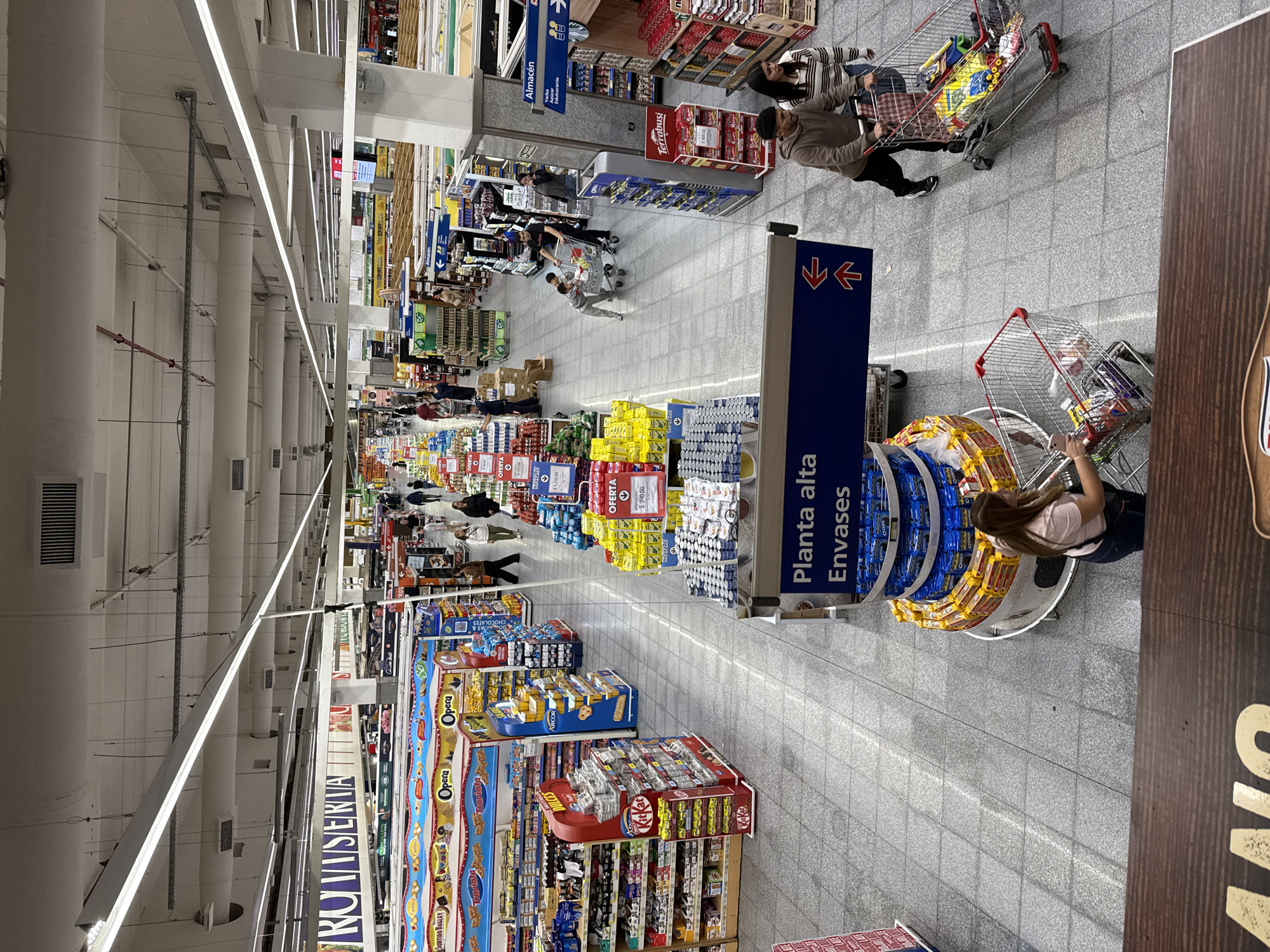 The image captures a wide-angle view of a brightly lit, long aisle within a large supermarket or warehouse store in Buenos Aires, Argentina. The setting features high ceilings with continuous fluorescent lighting strips and a grey tiled floor.
Numerous shoppers are present, pushing metal shopping carts filled with various items, or browsing the extensive product displays lining both sides of the aisle. People are casually dressed, indicating a typical shopping day.
The aisles are packed with a diverse range of packaged goods, snacks, and beverages. Prominently displayed products include cookies, crackers, chocolates, and other confectionery from brands such as "Terrabusi," "Arcor," "Opera," "Traviata," "Surtido Bagley," and "KitKat." There are also large stacks of what appear to be canned or bottled drinks, and packaged snacks.
Several signs with Spanish text are visible, providing directional and product information. A large blue sign with arrows points to "Planta alta Envases" (Upper floor Packaging/Containers). Another sign for a section called "Almacén" lists "Yerba Azúcar Edulcorante" (Yerba mate, Sugar, Sweetener), highlighting common Argentine staples. "OFERTA" (Offer/Sale) signs with prices, such as "$105.00," "$340.00," and "$5.240.00," are placed above certain product displays. The word "ROMISERTA" is also visible on one of the aisle markers. The overall atmosphere is busy and functional, characteristic of a modern grocery retail environment.