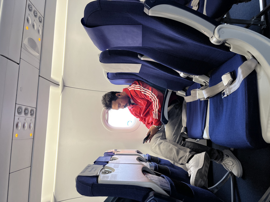 The image displays the interior of an airplane cabin, likely an economy class section, viewed from an aisle seat. A man is seated in a window seat on the right side of the frame. He is wearing a distinctive red jacket with white stripes along the sleeves, grey trousers, and white athletic sneakers. His head is bowed, and he is intently focused on a black smartphone he holds in his hands, suggesting he is texting, browsing, or engaged with its content.
The cabin features rows of blue fabric seats with grey and white plastic armrests and frames. Light-colored seatbelts, with the word "LIFT" visible on their buckles, are draped over the unoccupied seats both in front of and behind the man. In the foreground, two closed tray tables are attached to the back of the seats. Partial text such as "TO LOCK" and "DOWN" can be seen on their mechanisms, and a white safety information card is tucked into a slot on one.
A bright airplane window is positioned next to the man, allowing ample daylight to illuminate the cabin. To the left, along the ceiling, an overhead passenger service unit is visible. It features various controls including reading lights, some of which are indicated by small orange glows and light bulb icons, air vents, and call buttons marked with person icons. The cabin walls and ceiling are predominantly white, contributing to the bright interior. The man's concentrated posture suggests a moment of quiet activity or waiting during his journey, which is contextually related to Guwahati, India. No specific airline branding is identifiable within the frame.