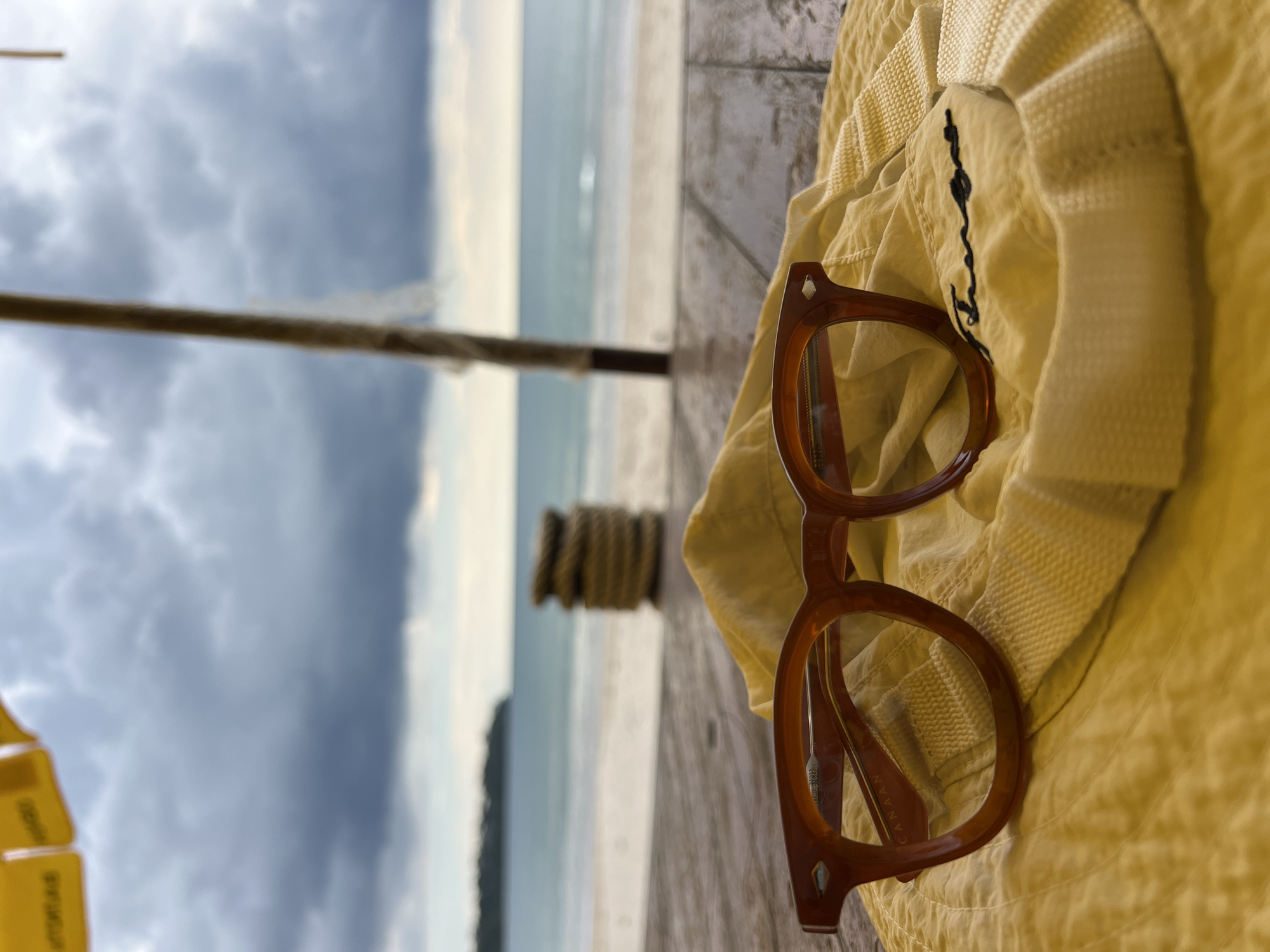 The image captures a scene on a beach, likely near a wooden deck or platform, on a cloudy day. A pair of brown-rimmed glasses rests on a light yellow hat. In the background, the beach stretches out, with the ocean meeting a stormy sky. A wooden post with coiled rope is visible. Part of a yellow umbrella is visible on the bottom left corner. The location of the photo is Unknown, Unknown.