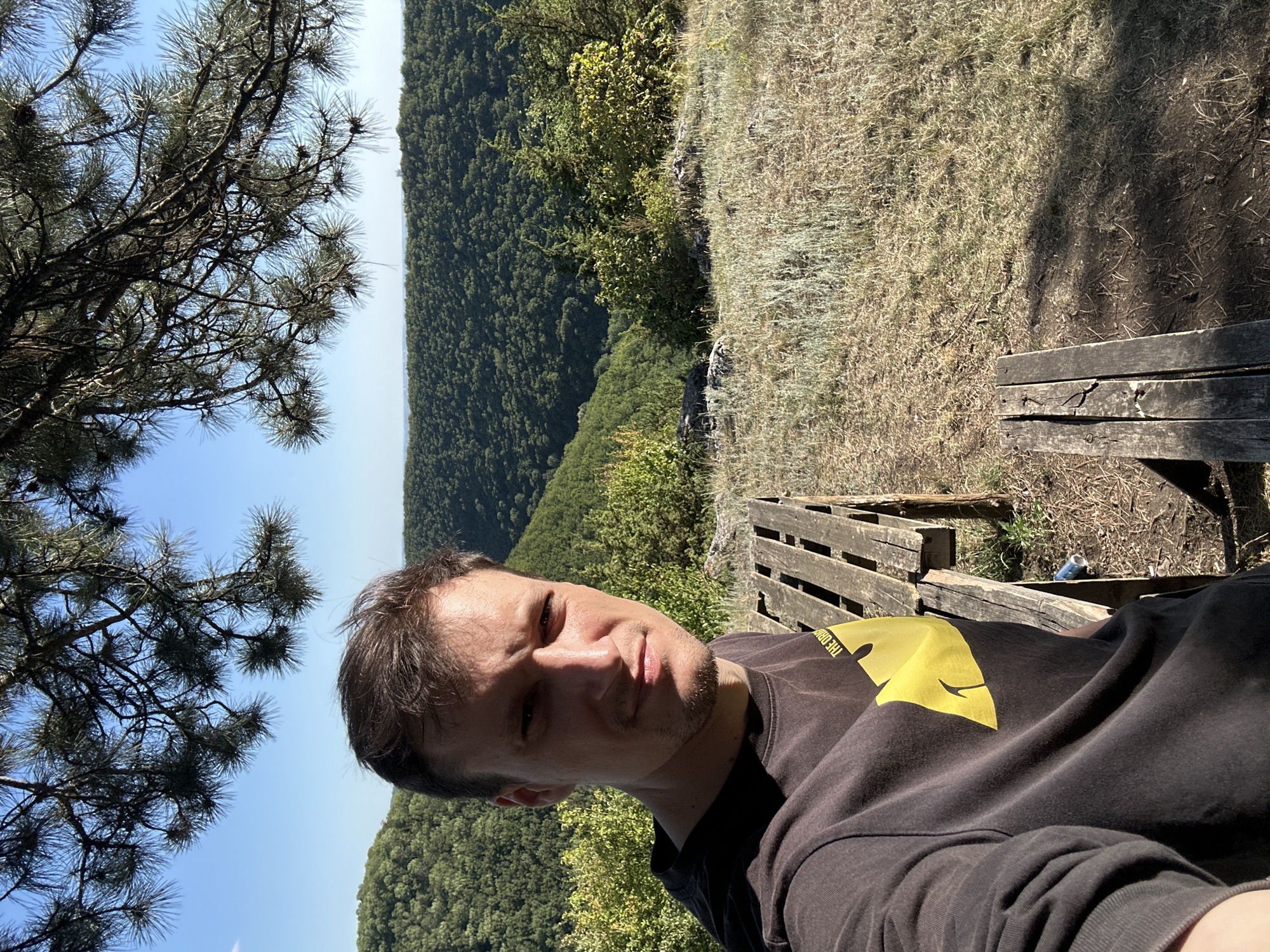 A man is taking a selfie outdoors with a lush, green forest in the background. He is wearing a dark sweatshirt with a bright yellow graphic. To his right, there is a wooden bench made from old planks. The scene is set on a hillside with dry grass and a clear blue sky. The overall impression is one of a peaceful day spent enjoying nature, possibly in a rural or park setting. The man's facial expression appears relaxed, suggesting he is enjoying the moment. The image captures a close-up of the man and the immediate surroundings, with the vast forest and sky filling the rest of the frame. There is a blue can visible on the ground behind the bench. The image appears to be taken during the daytime, with the sun casting shadows on the ground.