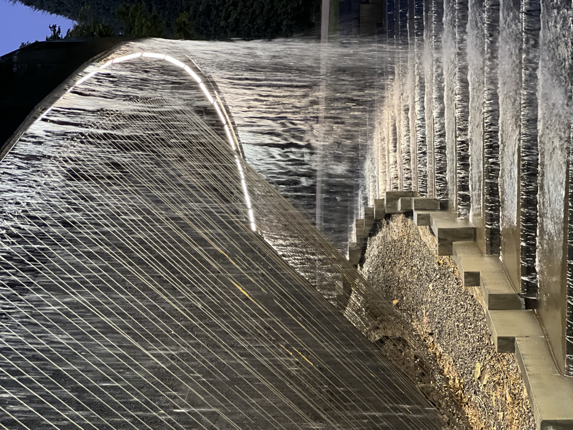 The image depicts a modern tiered water feature with water cascading down a series of concrete steps. A curved, illuminated band runs along the top edge of the structure from which the water falls. The background shows lush green trees and a dark blue sky, suggesting it is evening or twilight. The water feature is constructed with a dark, textured wall behind it, overlaid with numerous parallel, thin metal rods or wires. The scene appears to be an outdoor public space, possibly a park or plaza, designed for aesthetic appeal and relaxation. There are no people or animals visible in the image. The location is not specified as Krasnodar, Russia.