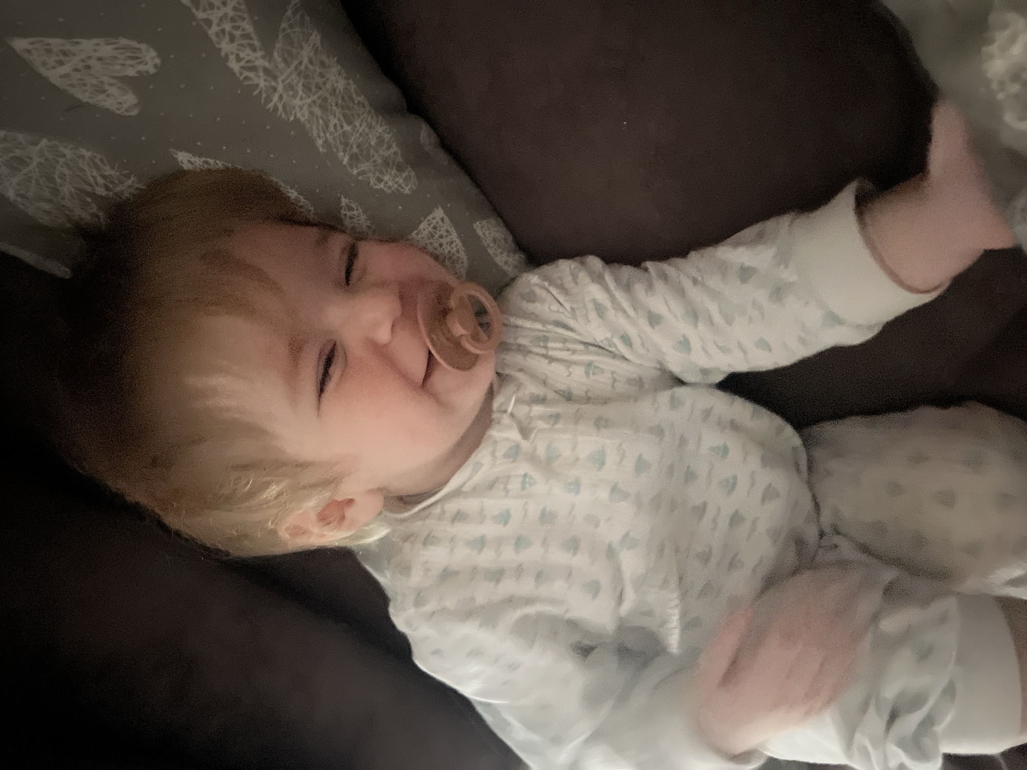 A baby with blond hair is lying on a dark brown surface, possibly a couch or bed. The baby has a pacifier in its mouth and is wearing a light-colored pajama set with a pattern of small blue shapes. The baby is resting on its back with its arms outstretched. To the upper left of the baby is a pillow with a light-colored pattern of abstract shapes resembling hearts. The scene is indoors and appears to be in a residential setting. There is no information in the image to connect it to the city of Noviy, Russia.