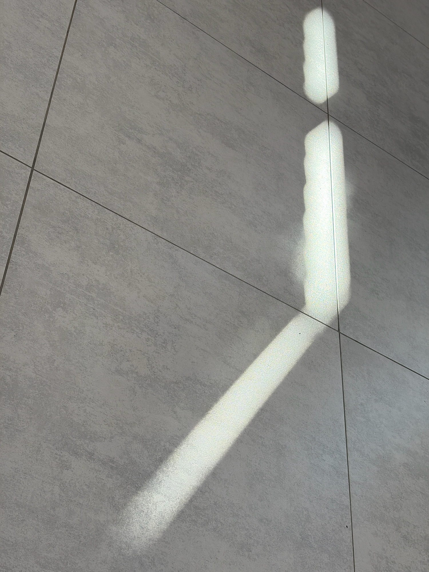 The image shows a close-up view of a tiled floor with a sunbeam casting a shadow in the shape of the lowercase letter "i". The tiles are light gray with a textured surface. The sunbeam is bright and casts a distinct shadow, with a slightly blurred outline. The shadow extends diagonally across the tiles, with the top of the "i" appearing as a short, illuminated rectangle, and the stem of the "i" extending further down the floor. The scene appears to be indoors, possibly in a home or public building, in Bogorodsk, Russia. There are no people or other objects visible in the image. The lighting suggests it is daytime.