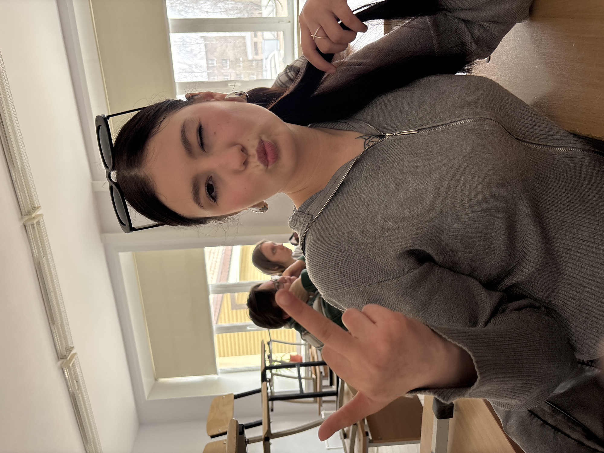 A young woman poses for a selfie in what appears to be a classroom in Chaikovsky, Russia. She is wearing a grey zip-up sweater and sunglasses perched on her head. She is winking, puckering her lips, and making a hand gesture, commonly referred to as the "rock on" gesture. She is holding her dark hair with a ring on her finger. A small tattoo is visible on her neck. Two other individuals are seated behind her. The classroom contains wooden chairs and tables. Windows with light-filtering blinds are in the background. The buildings visible through the windows suggest an urban environment.
