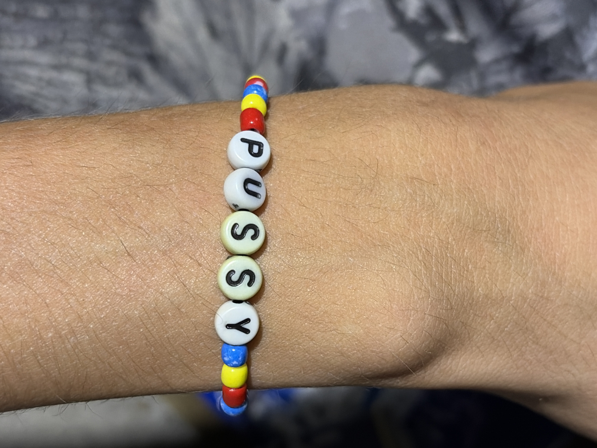 A close-up, eye-level shot shows a beaded bracelet on a person's arm. The bracelet is made of white letter beads spelling out "PUSSY" and is interspersed with colorful spacer beads in red, blue, and yellow. The bracelet is worn on a lightly tanned arm with visible hair follicles. The background is blurred, with hints of gray and blue fabric on the left and right sides, respectively. The scene appears to be indoors, but no specific location in Vratsa, Bulgaria can be determined from the visual information.