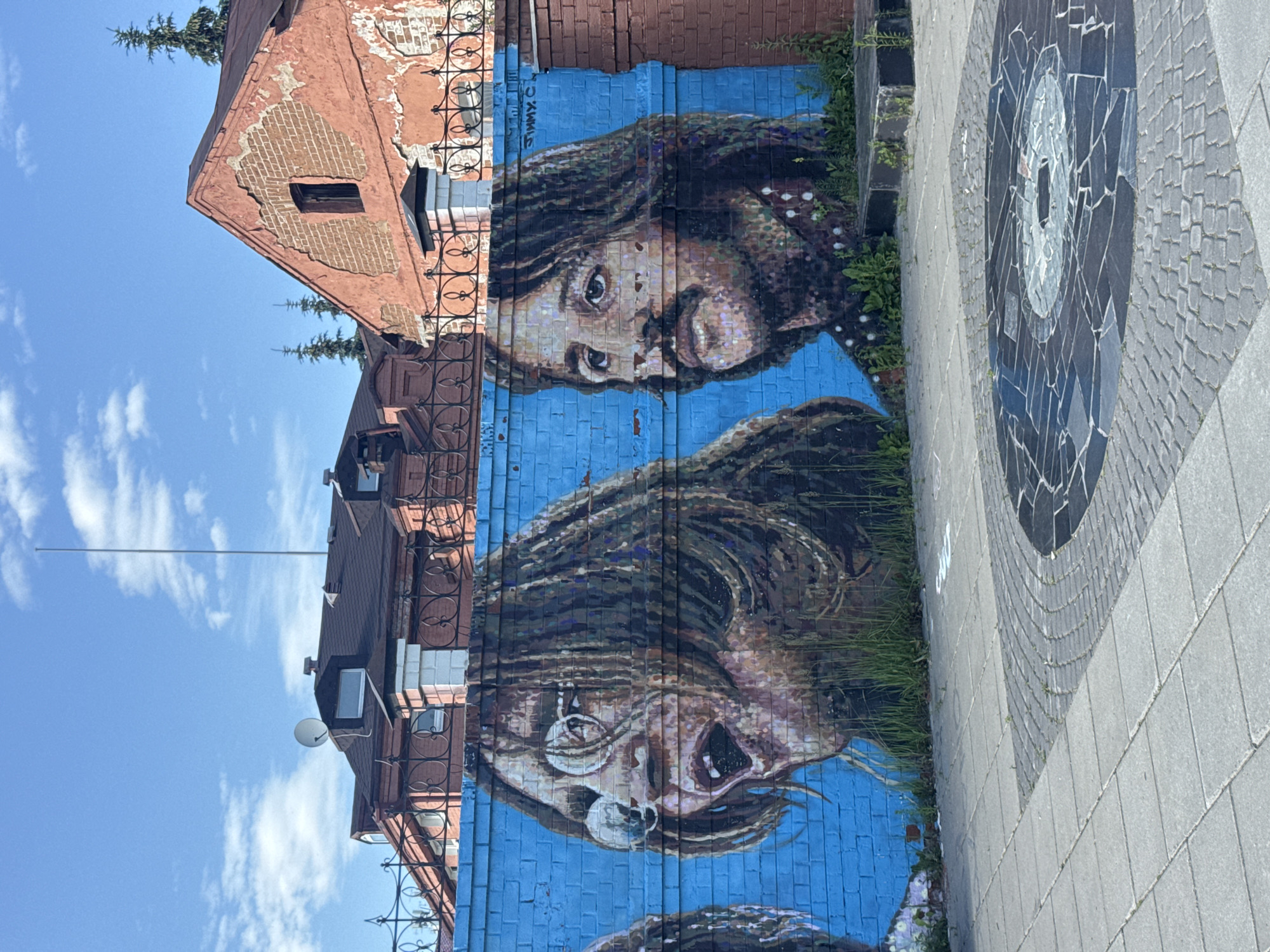 This image shows a mural of John Lennon and Paul McCartney painted on a bright blue brick wall in Yekaterinburg, Russia. The mural depicts the two musicians in a stylized manner, with John Lennon on the left wearing glasses and Paul McCartney on the right with a mustache. The wall is located in front of several buildings, including a reddish-orange brick building with peeling paint and a darker-roofed building behind a decorative black fence. In the foreground, a paved area features a circular mosaic design that resembles a vinyl record. Green grass and weeds grow along the base of the wall. The sky is blue with scattered white clouds.