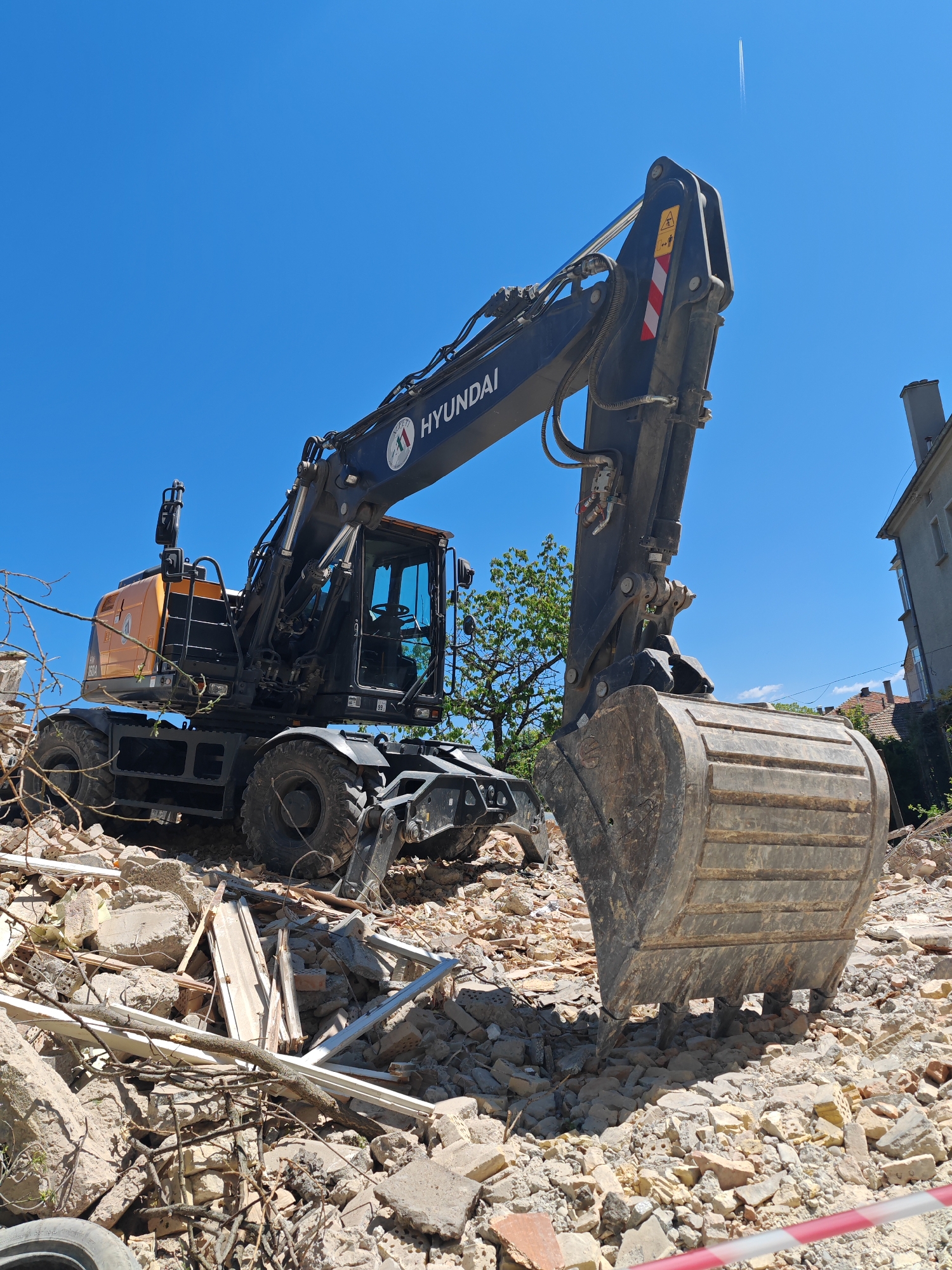 The image depicts a Hyundai excavator positioned atop a pile of rubble, likely from a demolished building. The rubble consists of brick, concrete, wood framing, and branches. A red and white striped barrier tape is visible in the foreground. The excavator is positioned with its bucket facing the viewer. The background features a clear blue sky and the corner of a building with a red tile roof. A contrail from an airplane can be seen in the top right corner of the image. The location is likely Shumen, Bulgaria, based on the identification mark on the machine.
