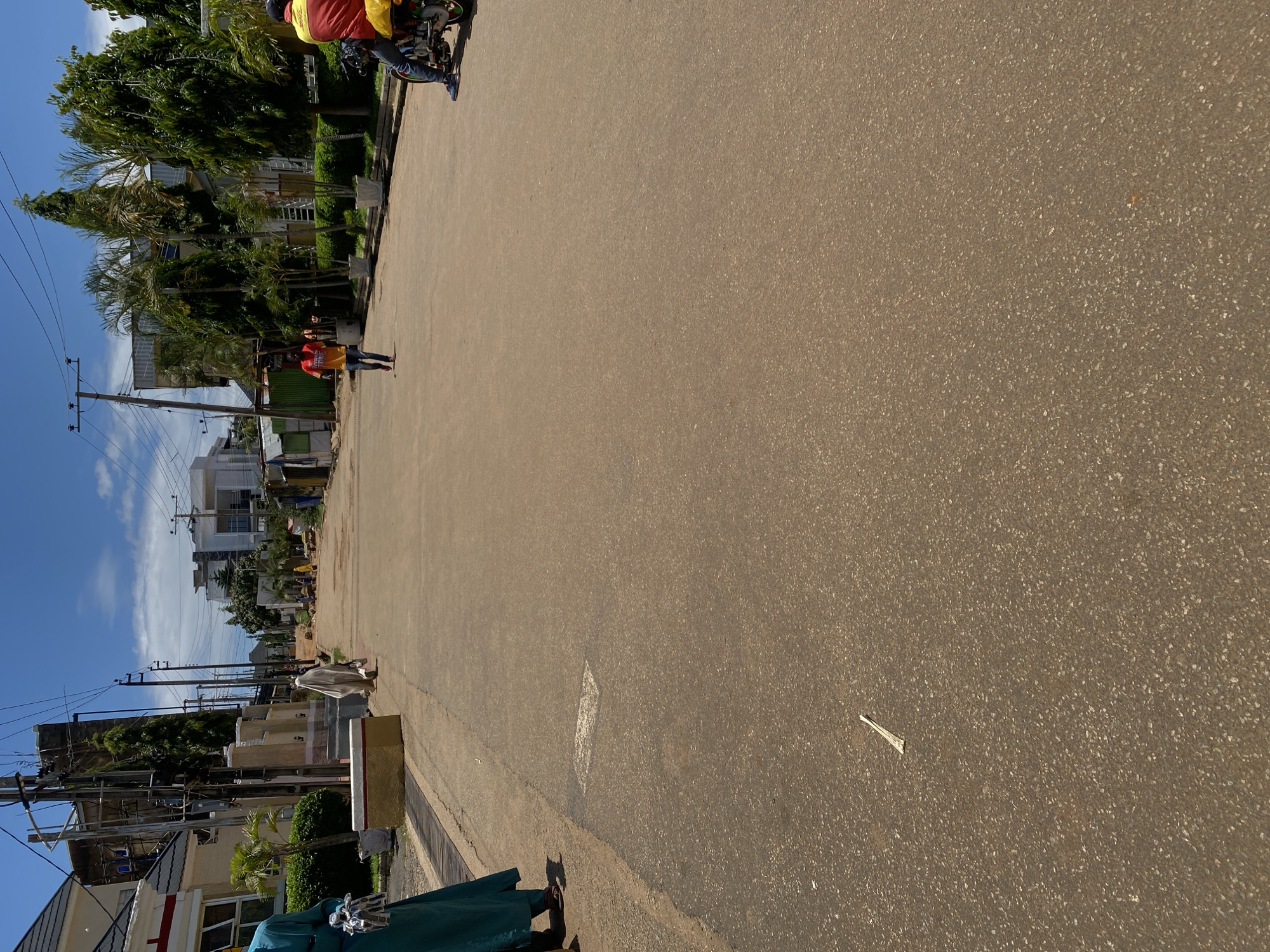 The image depicts a street scene in Jos, Nigeria, under a clear blue sky with scattered clouds. The road is paved with asphalt and appears to be moderately wide, with a gentle upward slope towards the background. On the left side of the road, a person in a teal-colored garment is partially visible, standing on a paved walkway adjacent to a building. Further down the road on the left, another individual in a white garment is walking away from the camera.
The right side of the road features a motorcycle with a rider wearing a red and yellow jacket. The rider is seated on the motorcycle, which is facing away from the camera. In the middle of the road, a person in a red shirt and blue jeans is walking towards the camera. The background reveals a mix of modern and older buildings, some with scaffolding, and lush green trees. Utility poles with overhead wires are prominent along the sides of the street. The overall impression is of a residential or commercial street in a developing urban area.