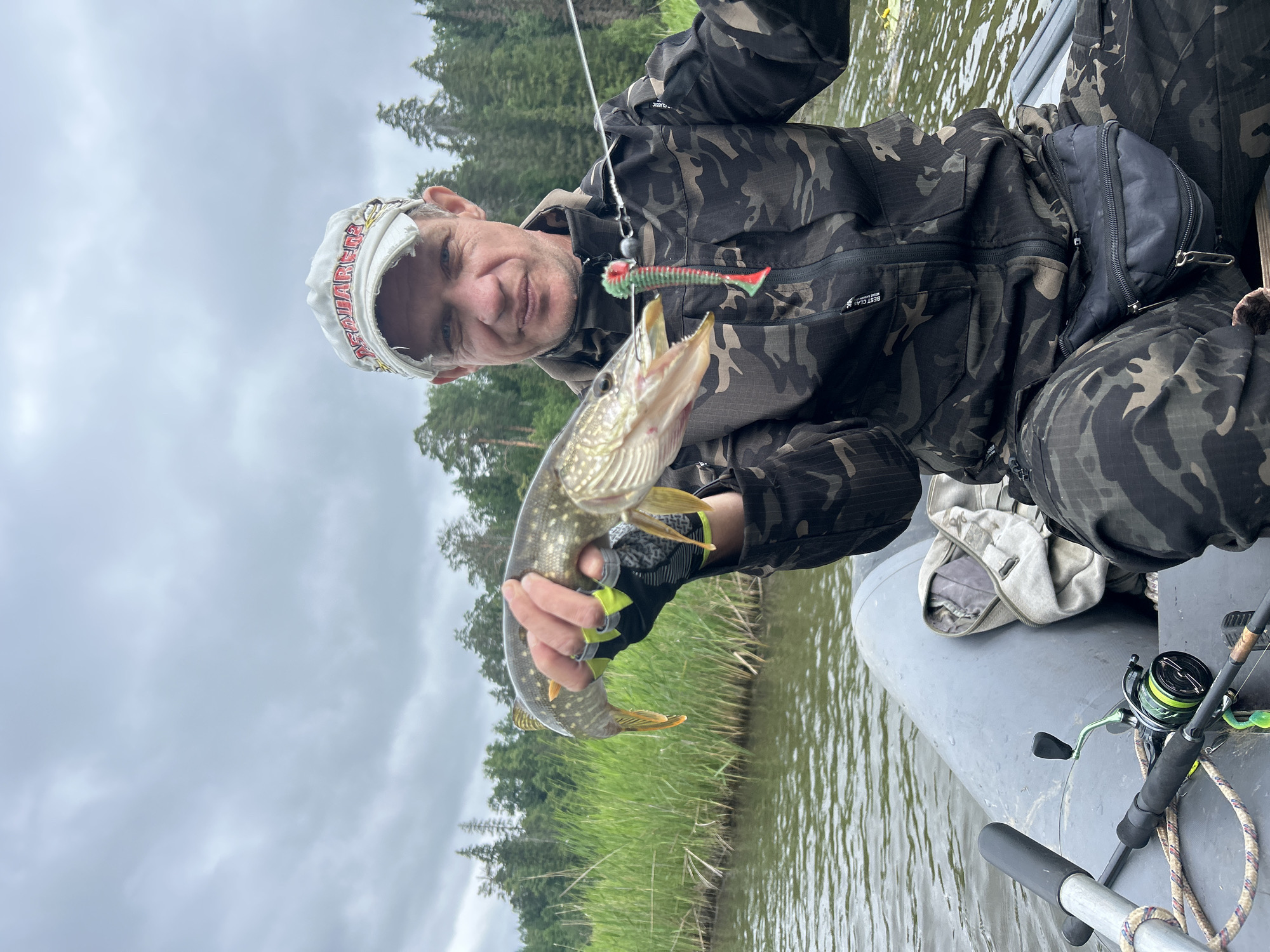 A man in camouflage clothing and a baseball cap sits in a grey inflatable boat on a body of water. He is holding a freshly caught pike, which is dangling from a fishing lure attached to a fishing line. The man is looking directly at the camera with his mouth slightly open. Behind him, tall green reeds line the shore, and a dense forest of trees stretches into the distance under a cloudy sky. On the boat floor, a fishing rod and reel are visible. The scene appears to be in a natural setting, likely a lake or river, in Detskii Dom (Bashenino), Russia.