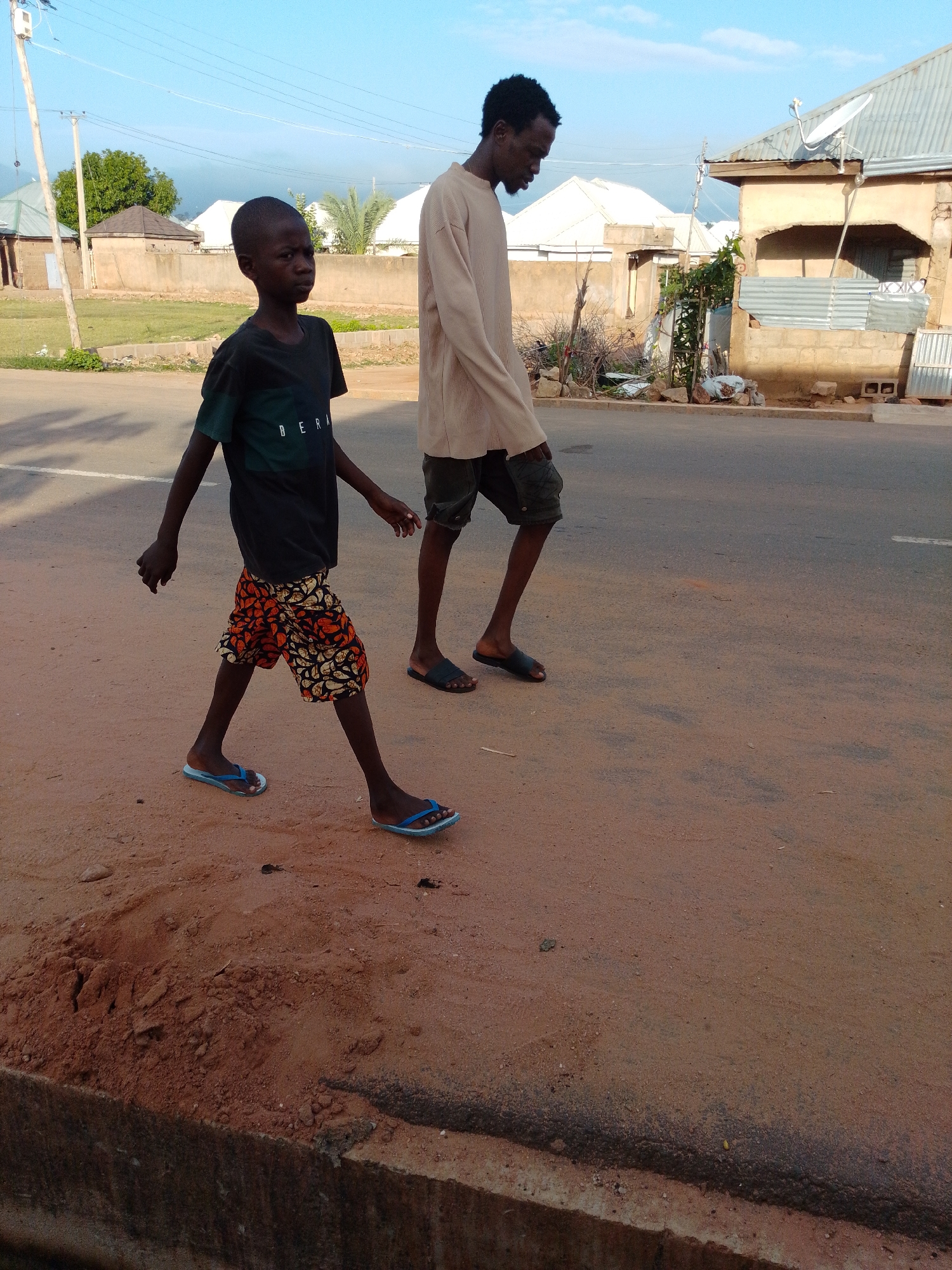 Two individuals, a child and a young adult male, are walking on a dirt and asphalt road. The child, in the foreground, is wearing a dark t-shirt with green lettering that reads "BERK" and patterned shorts. The young adult male, to the child's right and slightly behind, is wearing a long-sleeved, light-colored shirt and dark shorts. They are walking away from the camera, towards the right side of the frame. The environment appears to be a semi-urban setting in Gombe, Nigeria, with buildings and vegetation visible in the background under a clear blue sky. The road surface is a mix of reddish-brown dirt and darker asphalt.