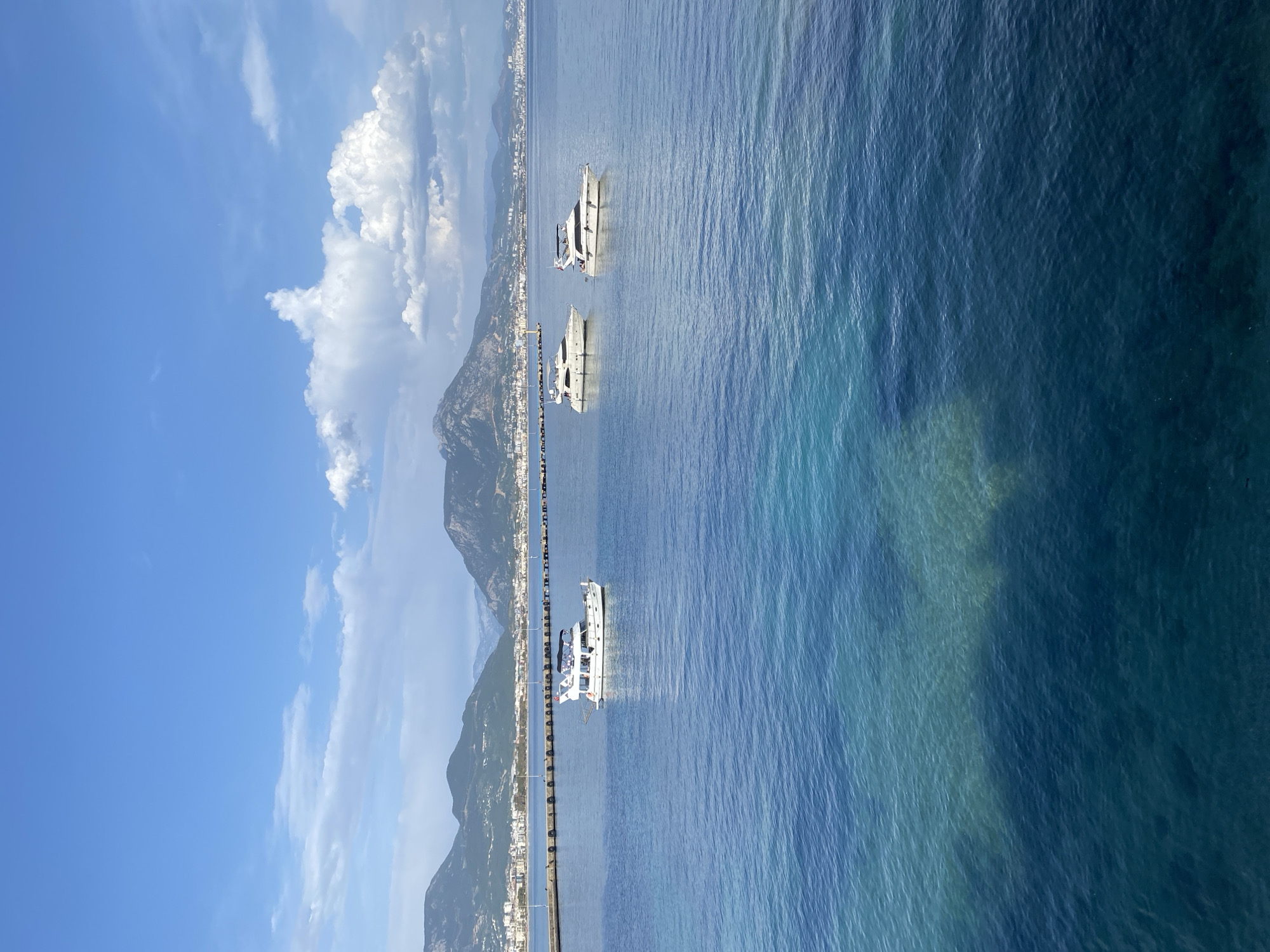 The image depicts a coastal scene in Alanya, Turkey. Three white yachts are anchored in the turquoise water near a long pier. The city stretches along the coastline at the base of a mountain range that rises prominently in the background. Cumulus clouds are visible in the blue sky above the mountain. The pier is lined with pylons.