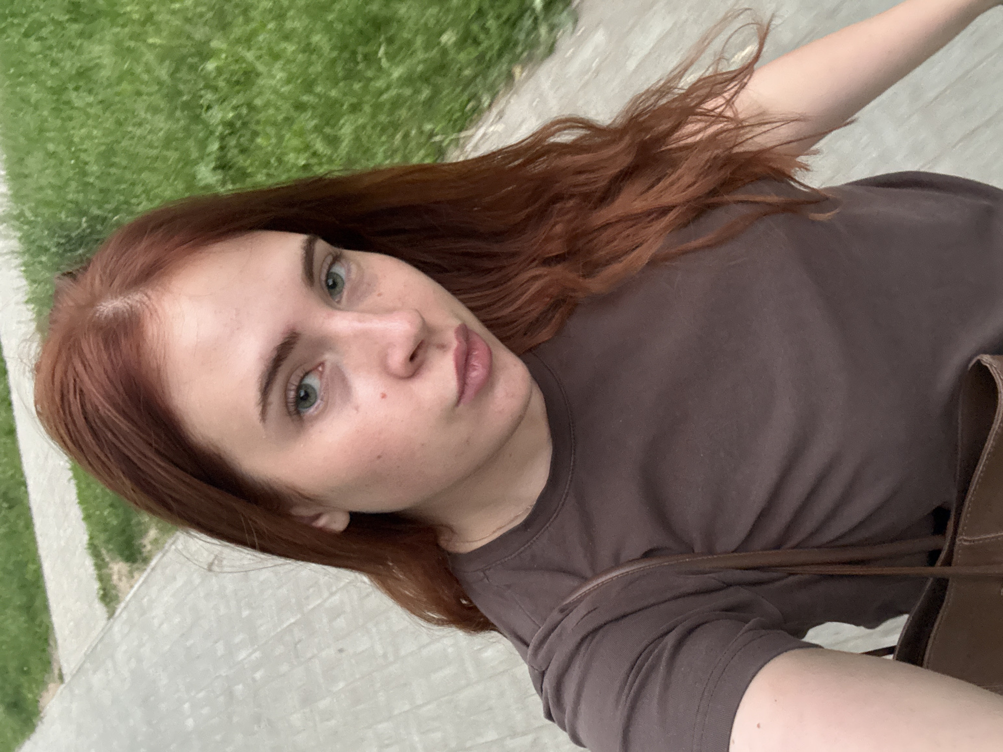 An overhead, medium shot captures a young Caucasian woman with long, wavy auburn hair and light green eyes, looking into the camera while walking on a paved path next to a grassy area. She is wearing a plain brown t-shirt and a brown purse is visible slung over her left shoulder. The scene appears to be outdoors, possibly in a park or residential area within the city of Chaikovsky, Russia. The lighting is natural and diffused, suggesting it may be overcast or late afternoon.
